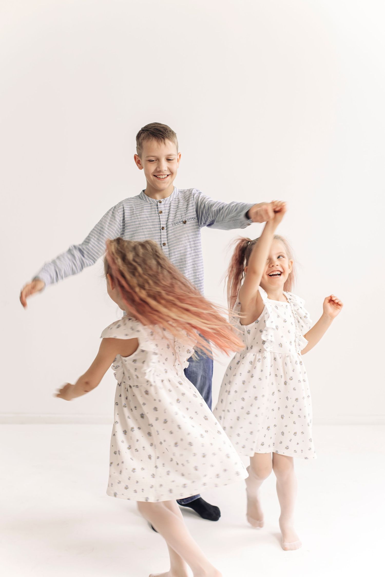 Three happy children dancing together, two girls in floral dresses and a boy in striped shirt, on white background.