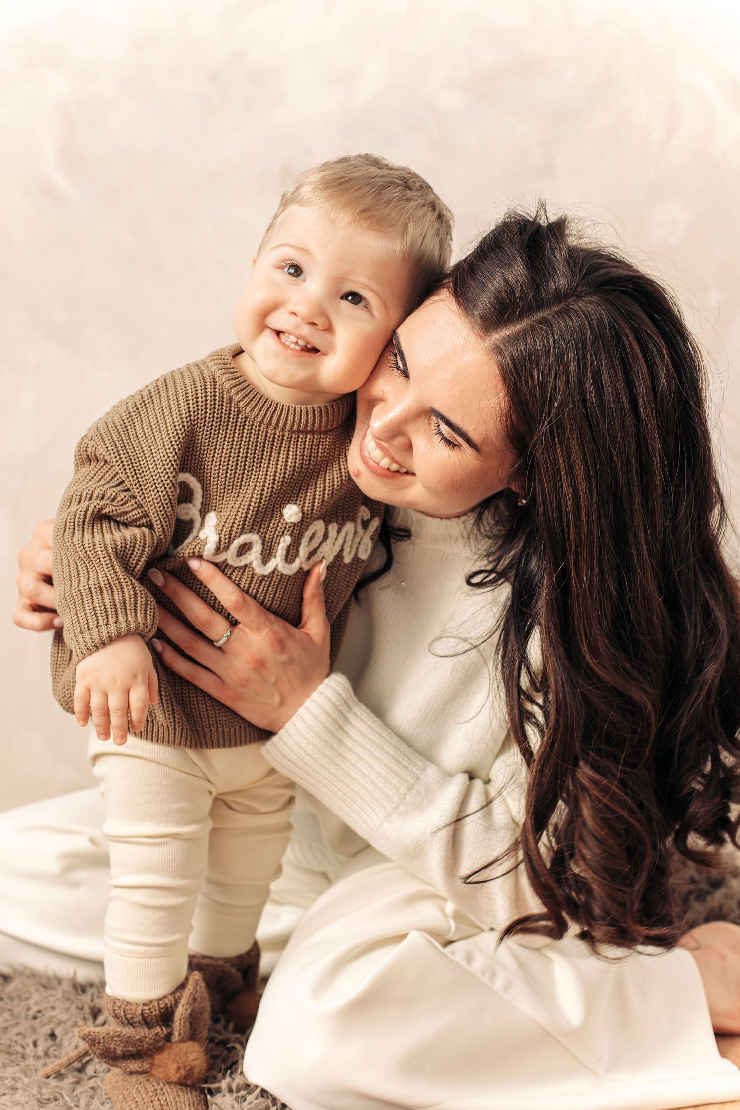 A smiling mother in a white sweater hugs her happy toddler in a brown knit sweater during a cozy studio photo session.