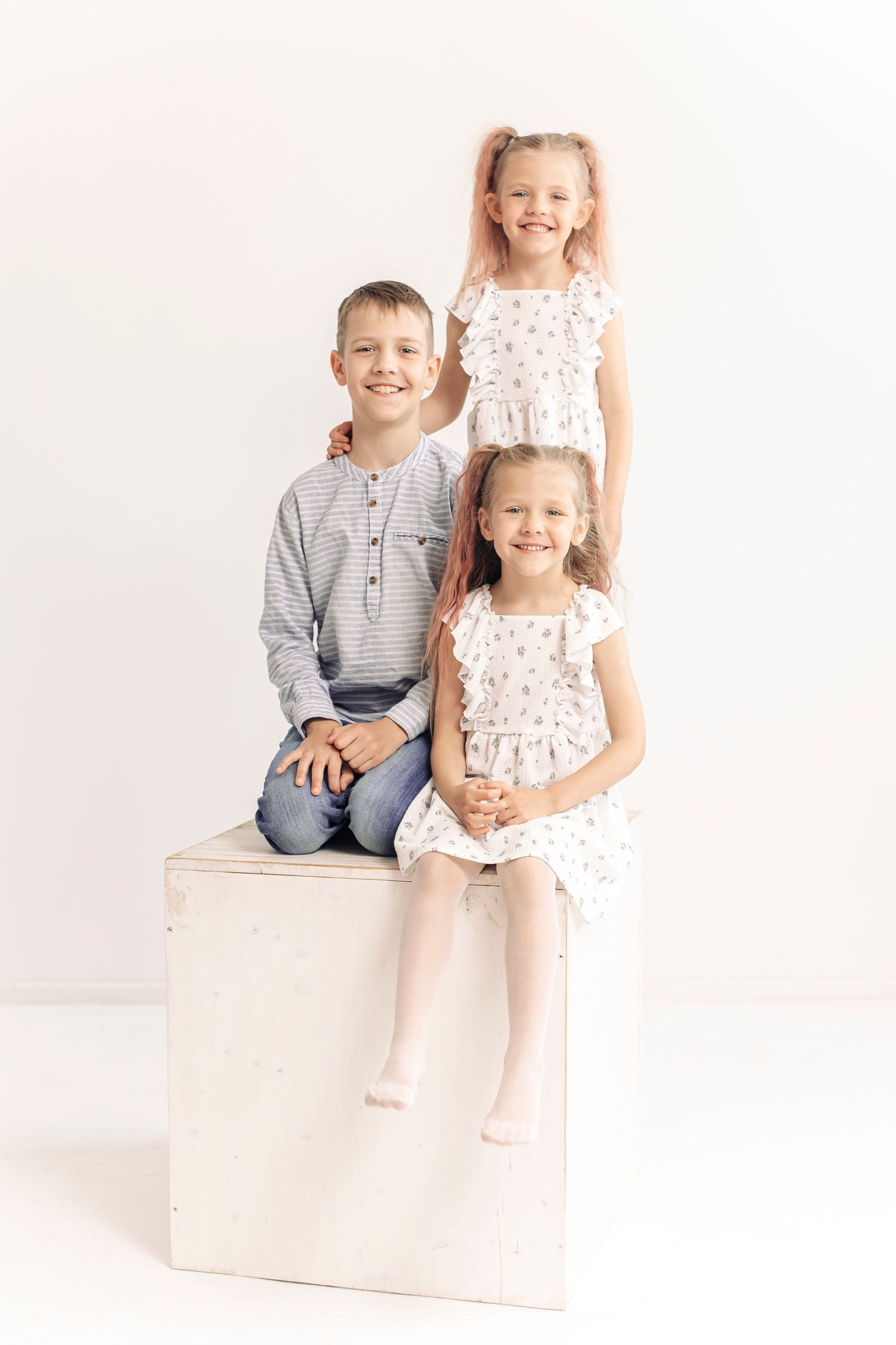 Three smiling children pose together on a white wooden cube against a light background in a studio portrait.