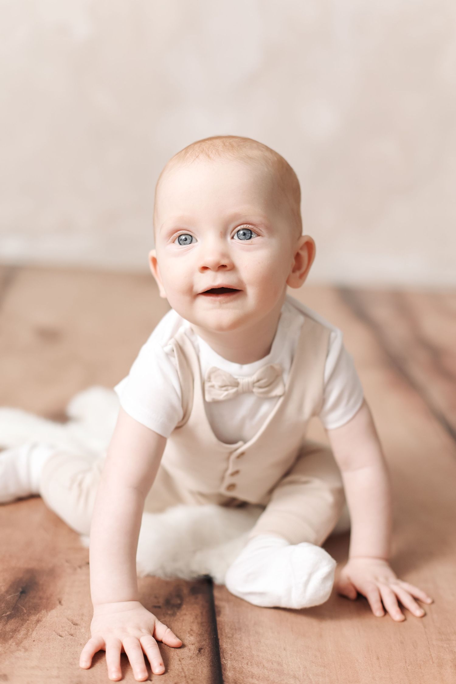Adorable baby in white outfit with bow tie sitting on wooden floor, looking up with bright blue eyes.
