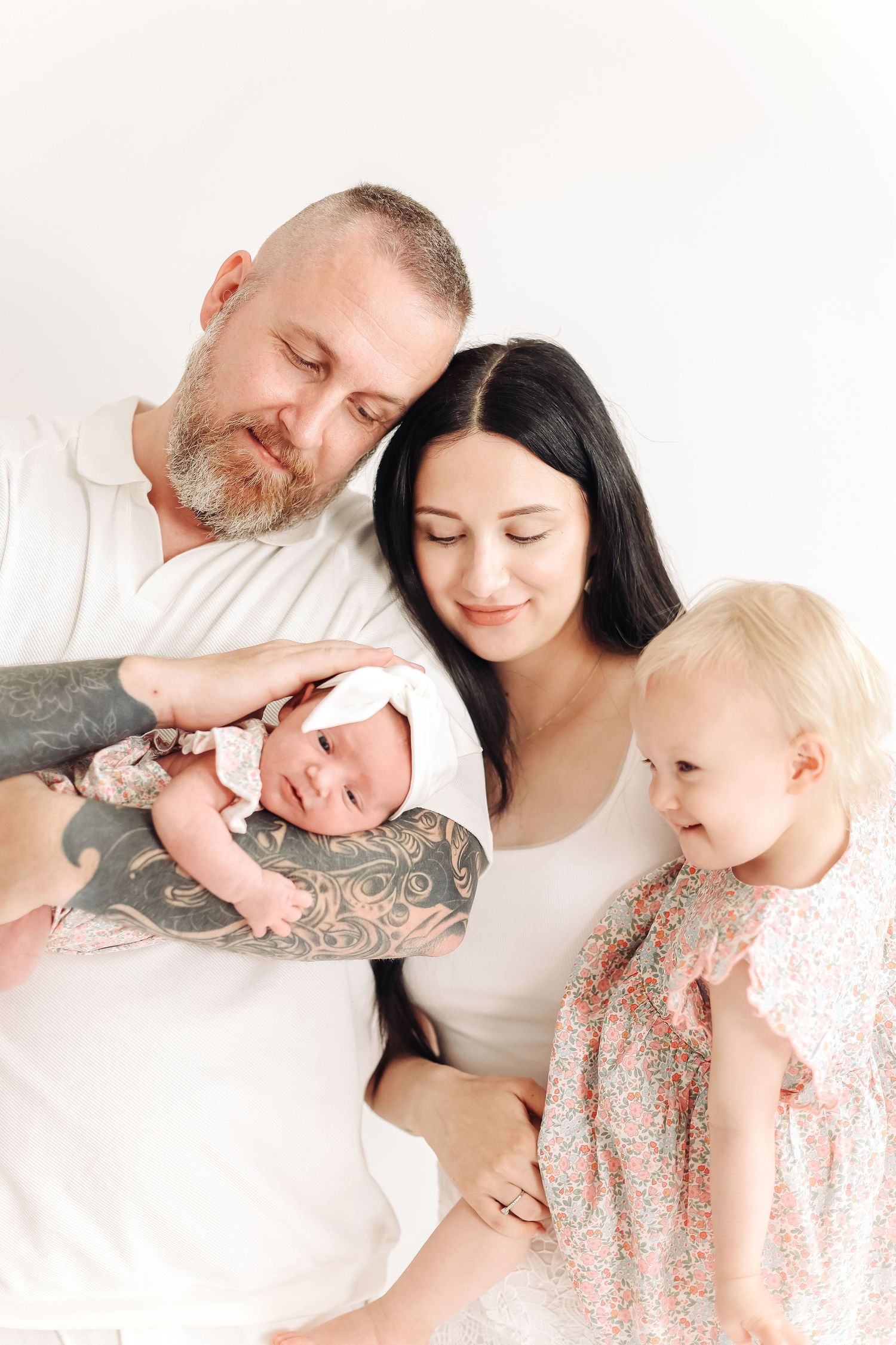A tattooed father and dark-haired mother lovingly hold their newborn while their toddler daughter looks on, all in white.