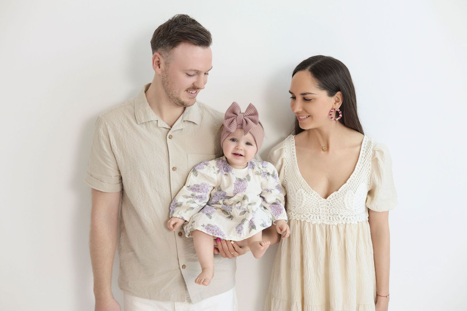A happy young family with a man, woman, and baby girl in a bow posing together against a white background.