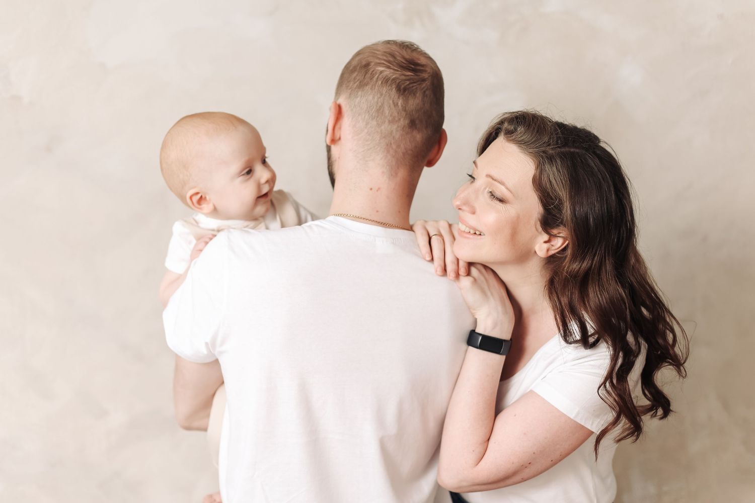 A young family with a mother, father, and baby posing together against a neutral beige background in white outfits.