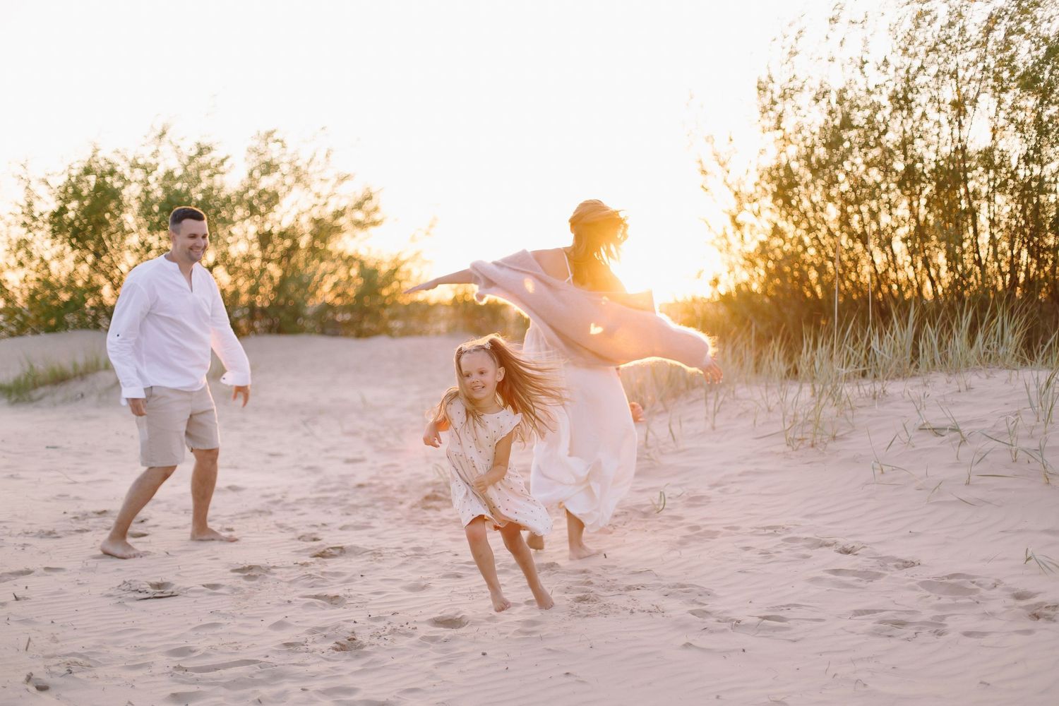 Family playing on sandy beach at golden sunset, parents and child laughing in white outfits with warm sunlight.