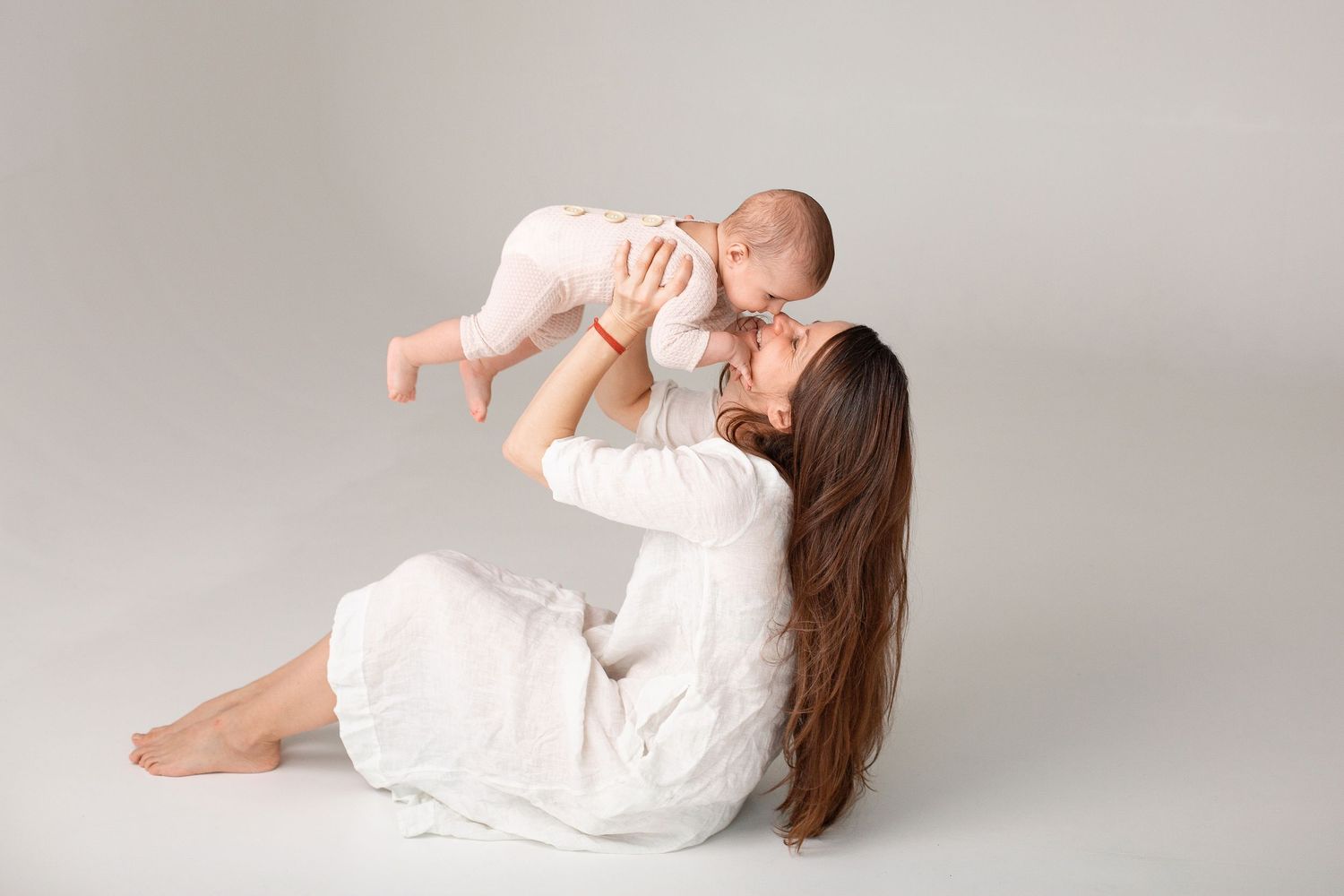 A mother in a white dress sits on the floor, lifting her smiling baby in a pink outfit up to kiss them. Māmiņdienas fotosesija Rīgā.