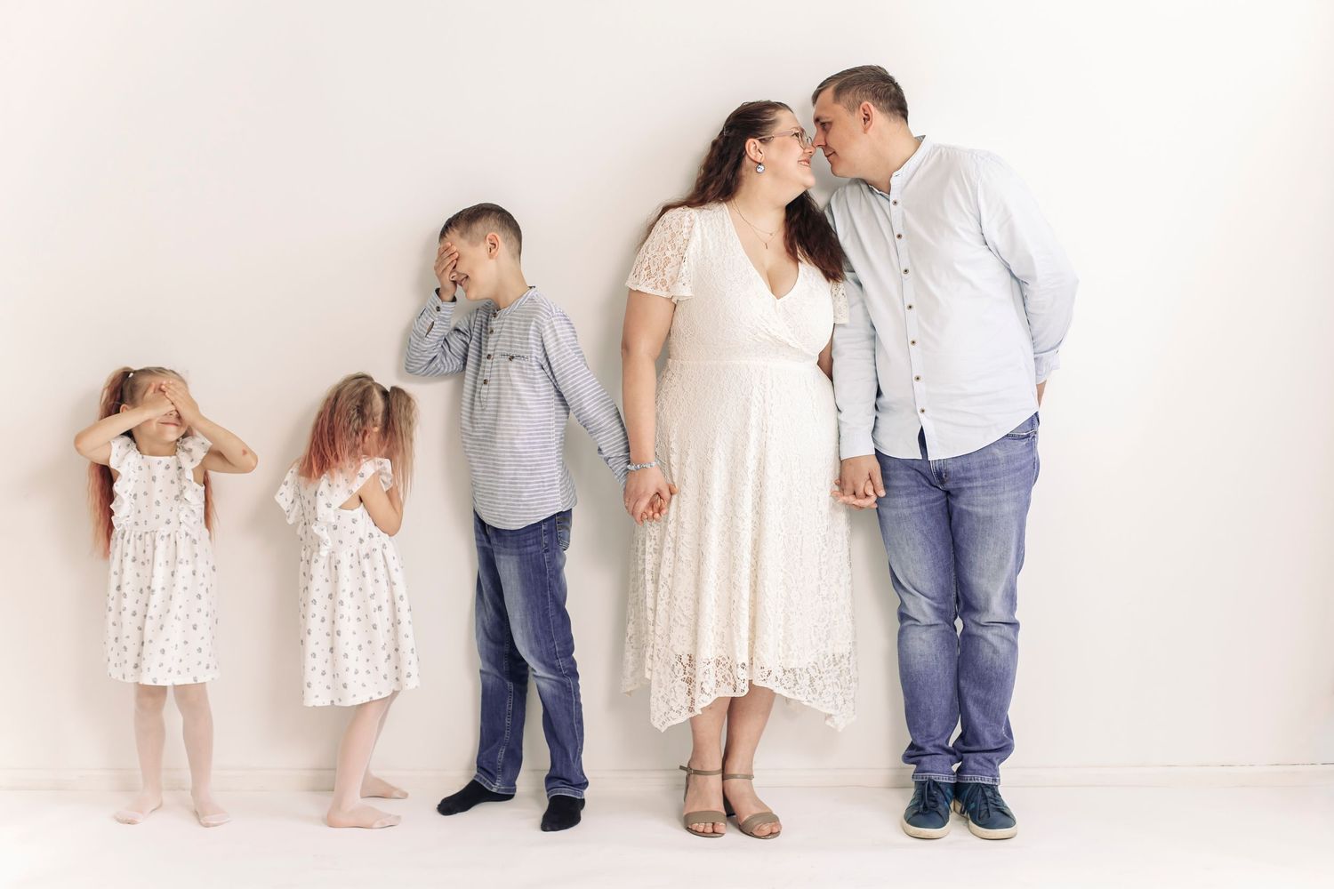 Family portrait with parents kissing and three children posing against a white wall in coordinated outfits.