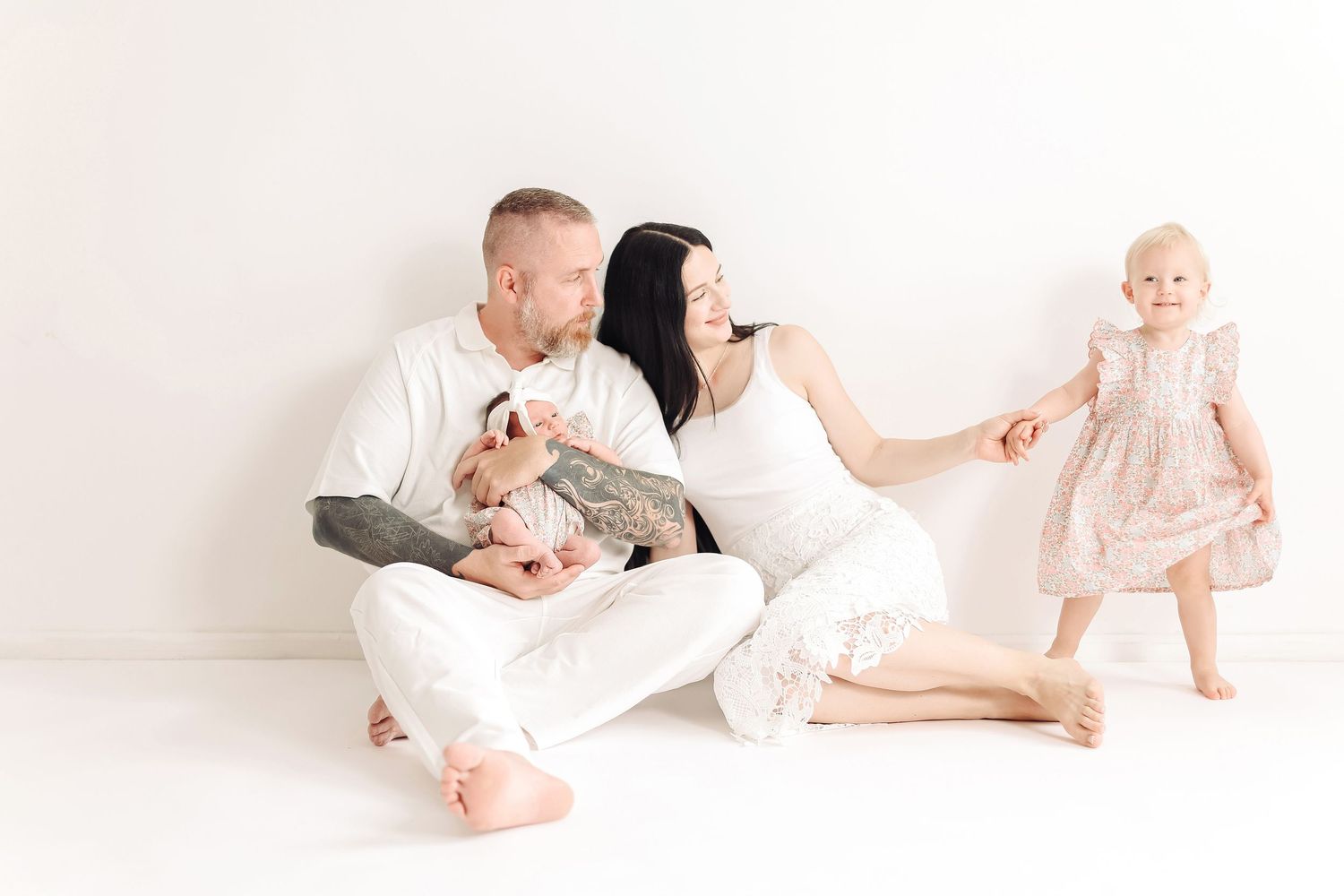 Family in white outfits sitting against white wall, parents holding newborn while toddler girl in pink dress stands nearby.