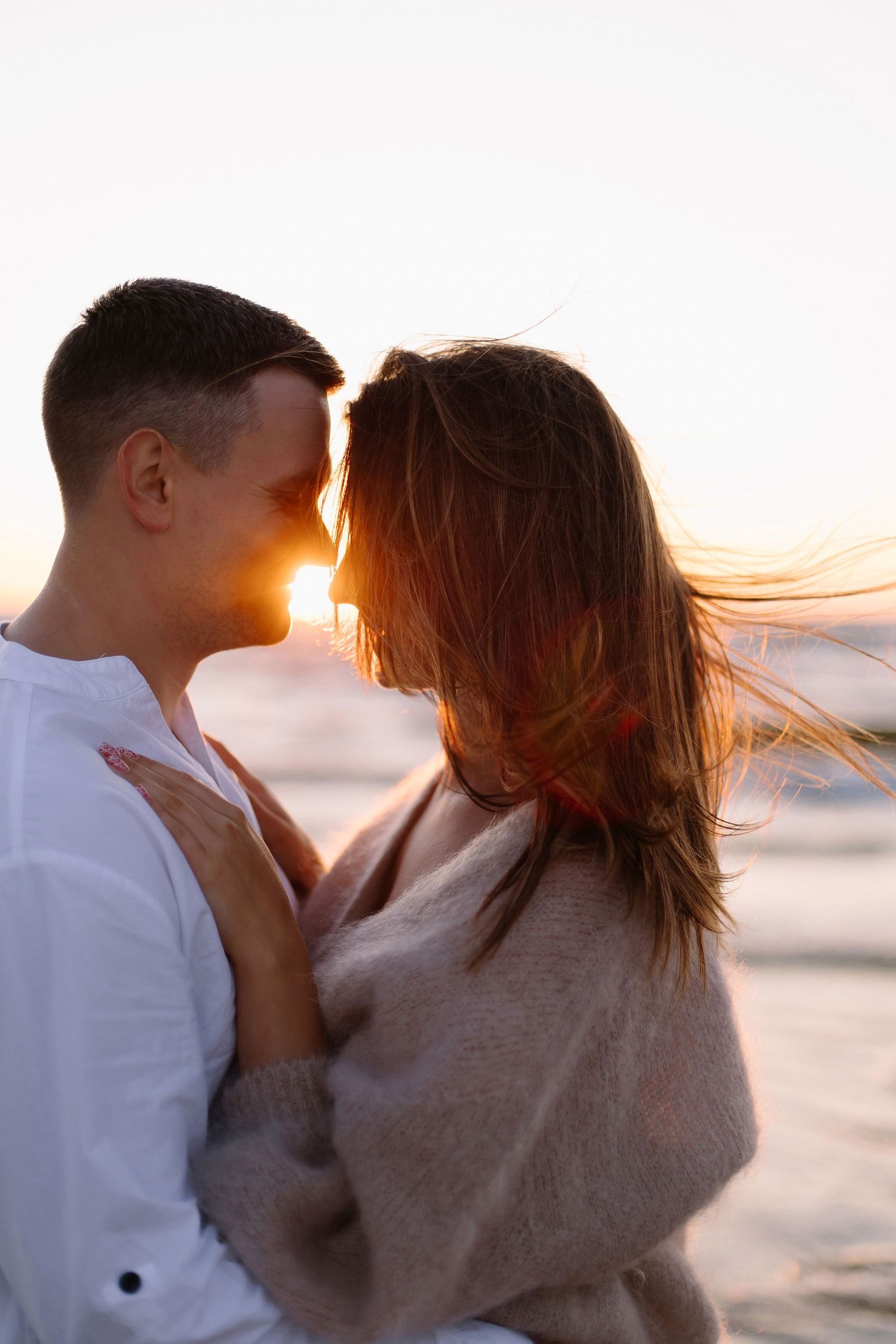 Romantic couple embracing at sunset on a beach, with golden light glowing between them and wind blowing her hair.