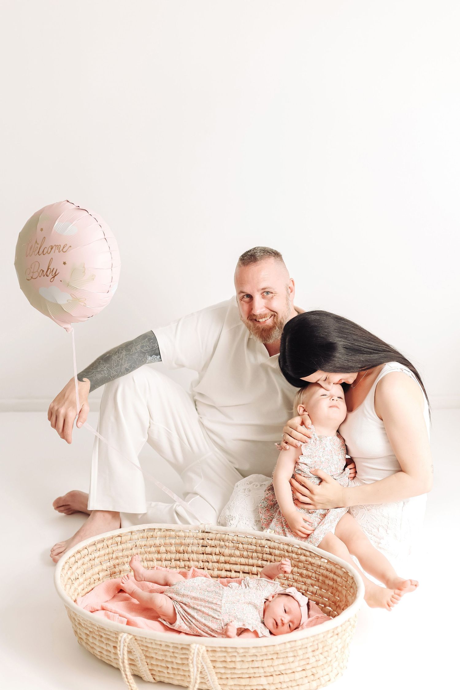 Happy family with newborn baby in basket, toddler, and pink balloon on white background.