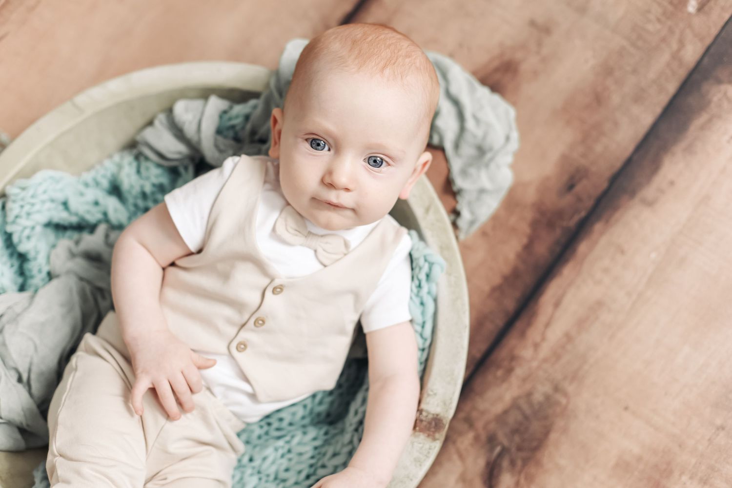 Adorable baby in beige outfit with bow tie resting in a round wooden bowl with teal knit blanket.