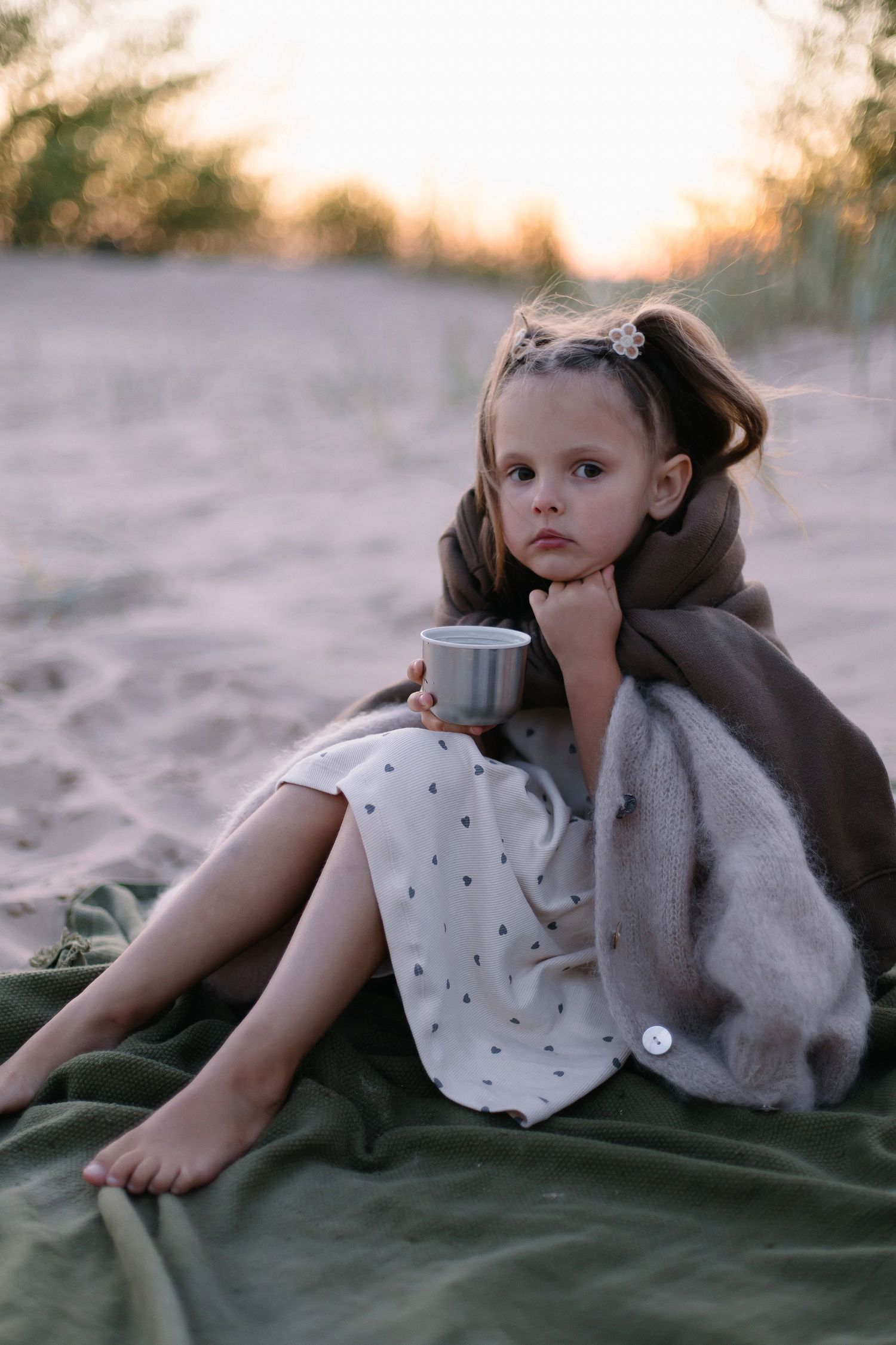 Young girl wrapped in blanket sitting on sandy beach at sunset, holding a small cup with a thoughtful expression.