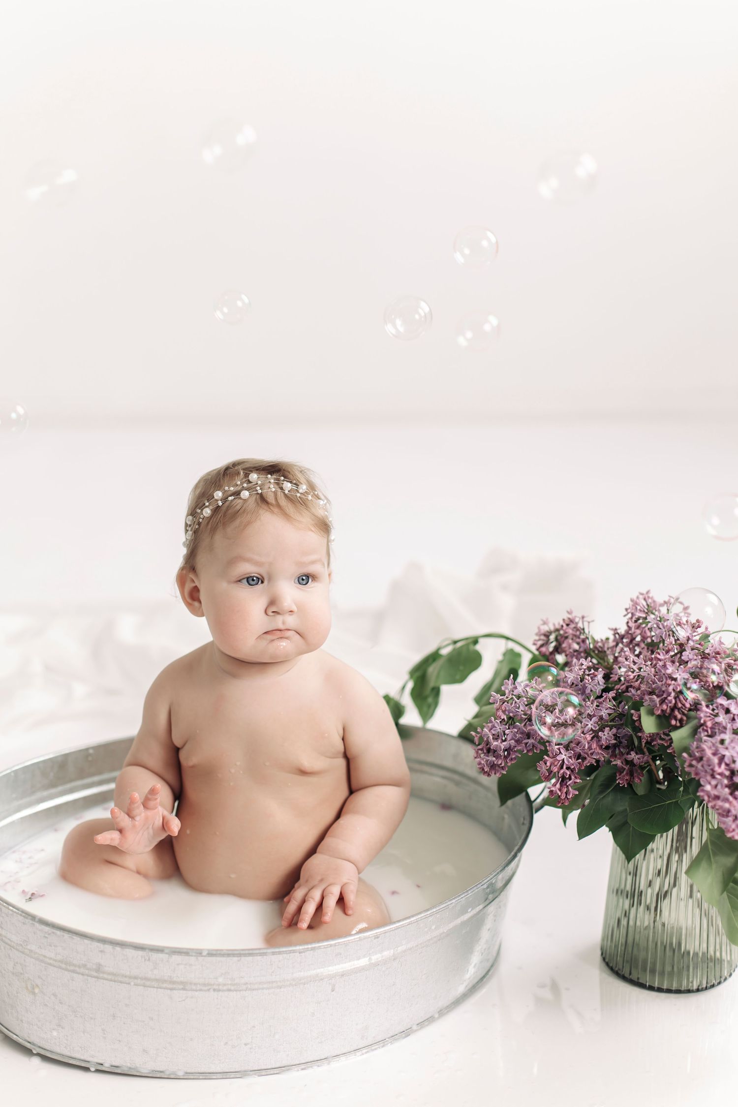 Baby girl in floral headband sits in vintage metal tub with milk bath, surrounded by bubbles and lilac flowers.