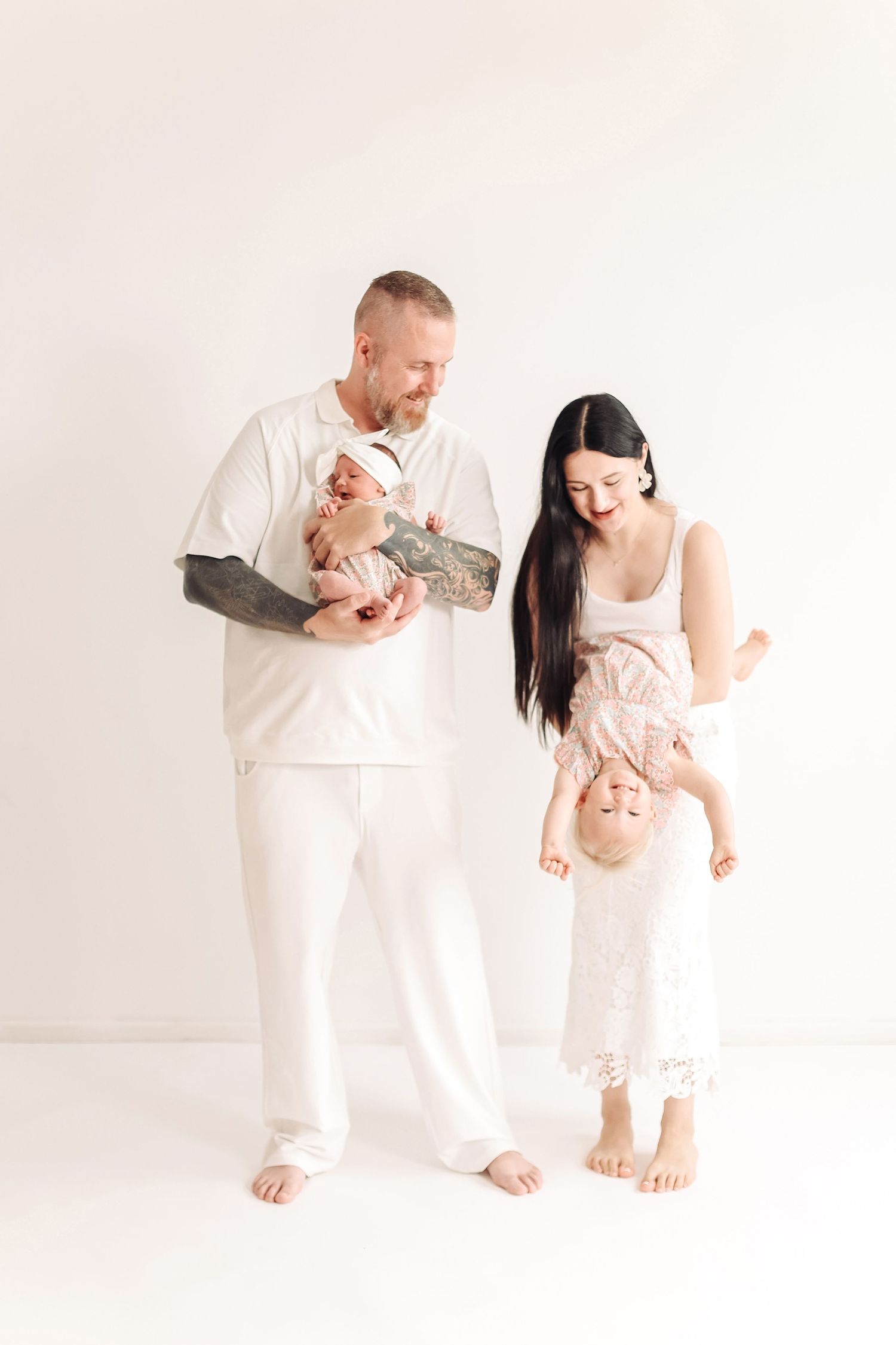 A tattooed father holds a newborn while mother playfully dangles their toddler upside down in a bright white studio.