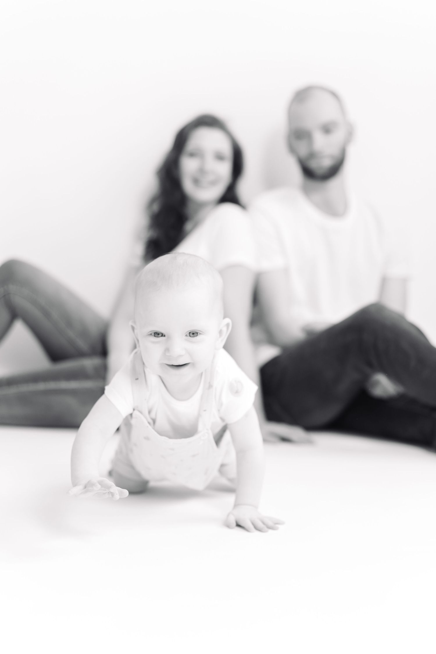 Black and white photo of a happy baby crawling toward camera with smiling parents sitting in background.