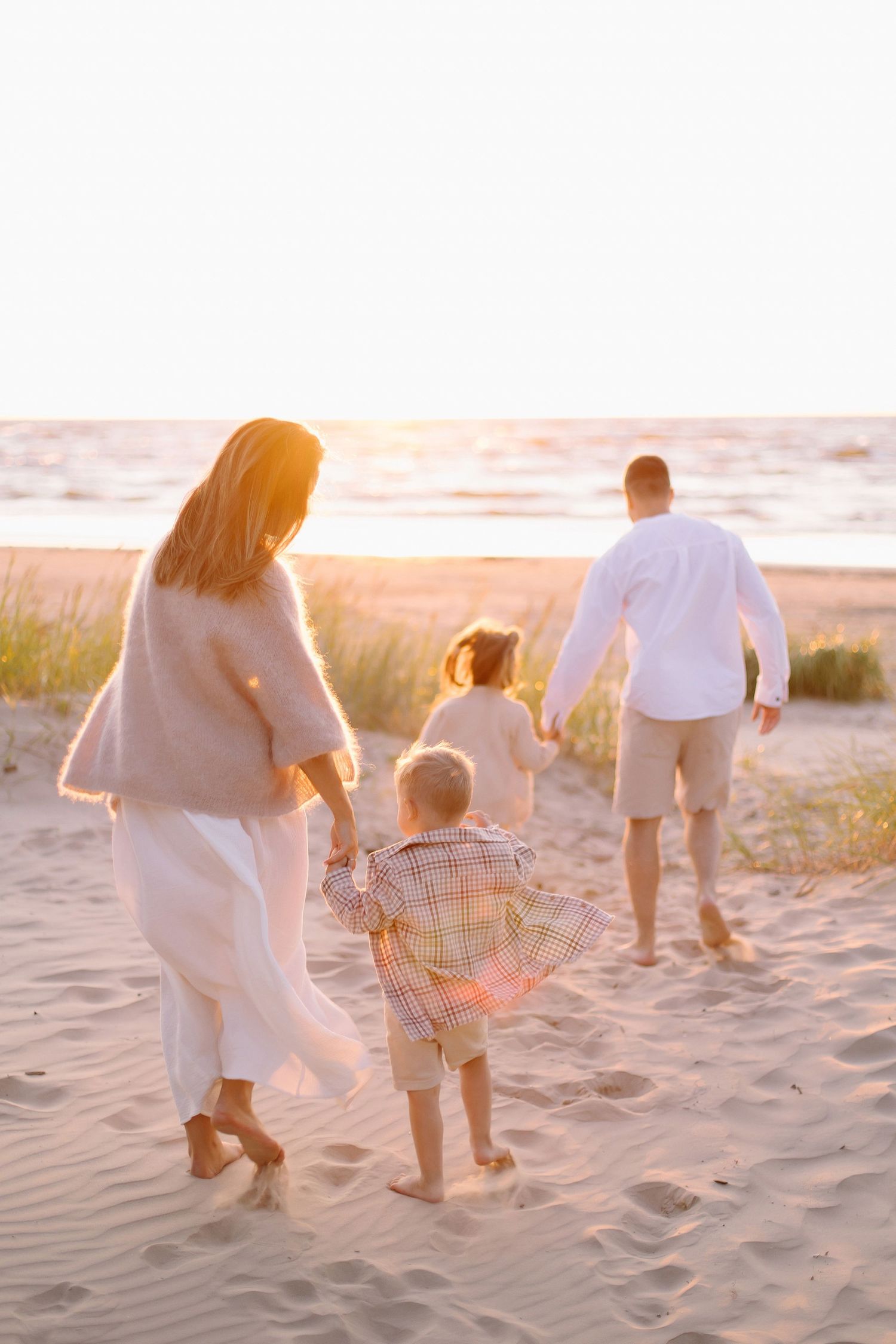 Family of four walking barefoot on sandy beach at golden sunset, parents holding children's hands.