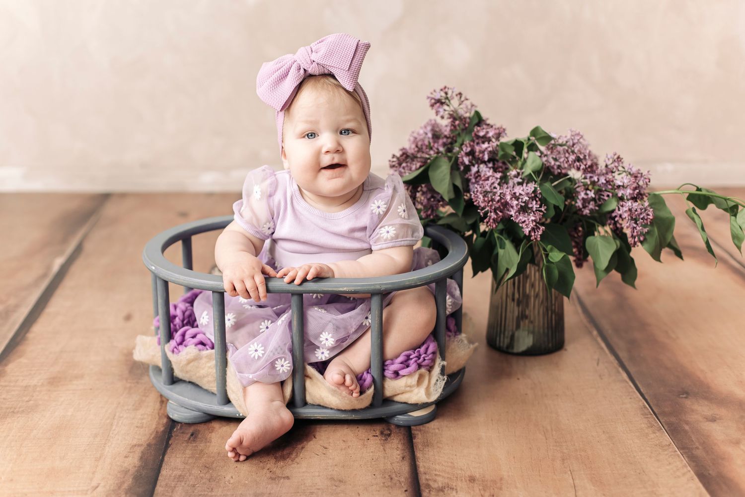 Smiling baby girl in purple outfit and bow headband sitting in wooden basket with lilac flowers on hardwood floor.