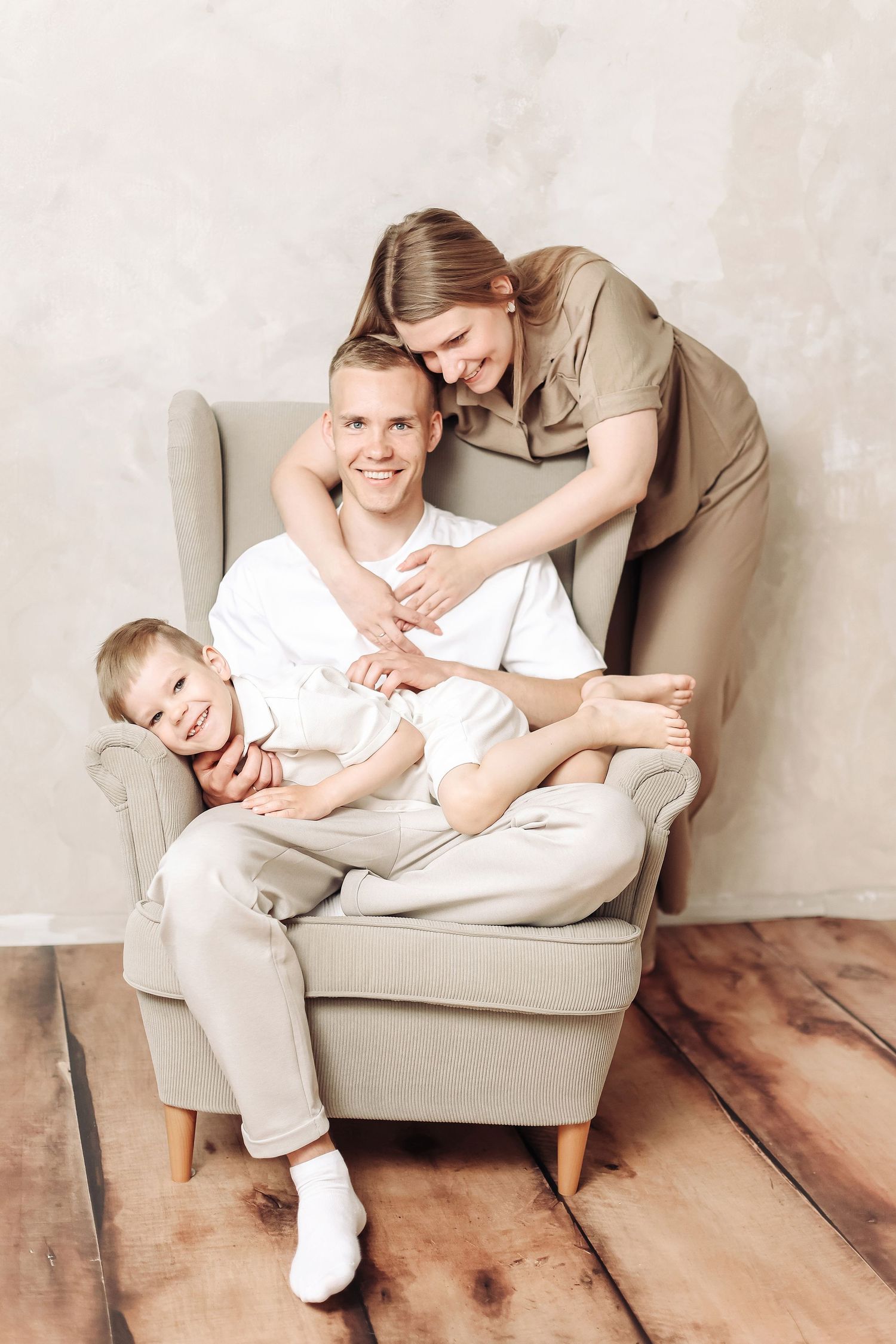 Happy family of three posing in a gray armchair, mother standing behind father and child sitting on his lap.