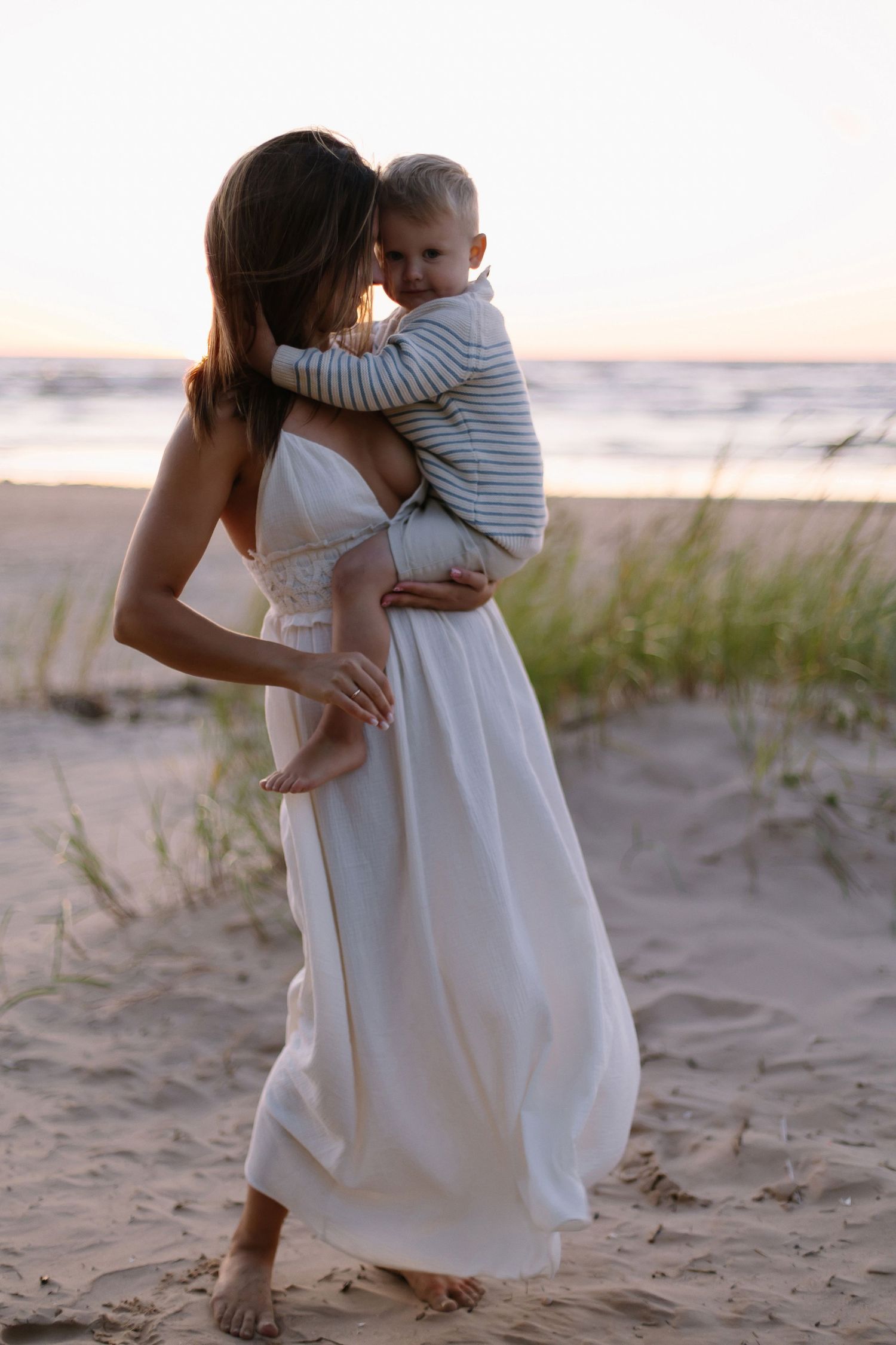 Mother in white dress holding baby on sandy beach at sunset, sharing tender moment.