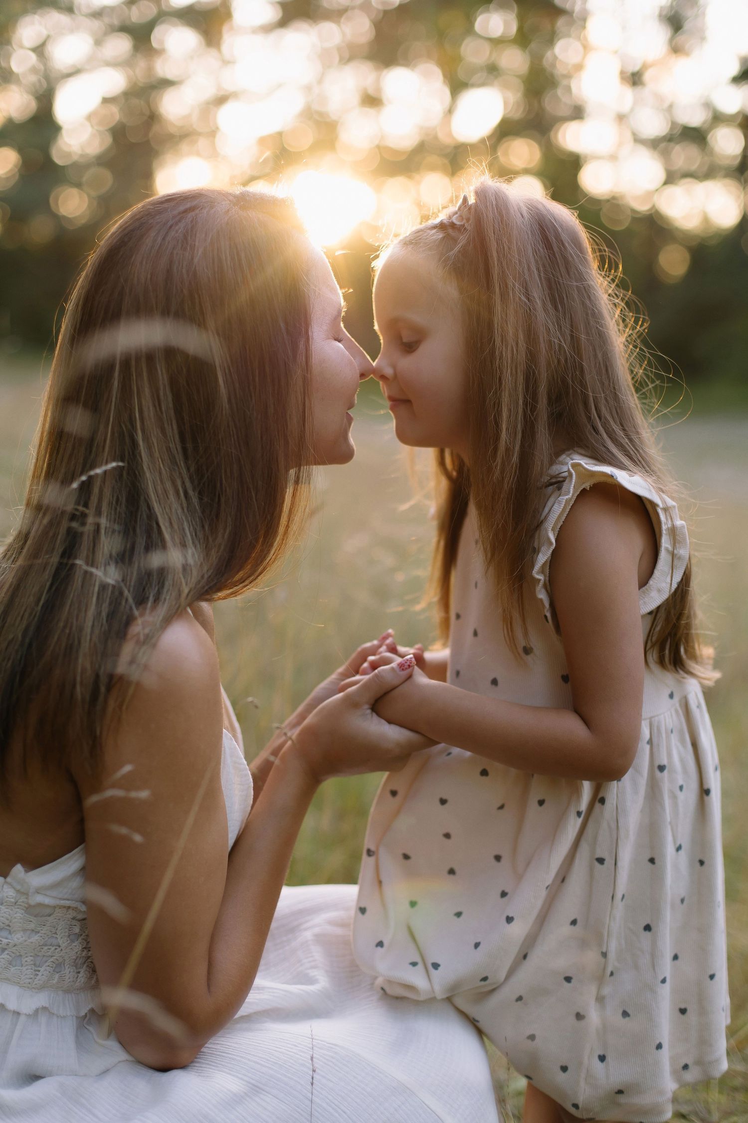 Mother and daughter share a tender nose kiss in golden sunset light, holding hands in a sunny meadow.