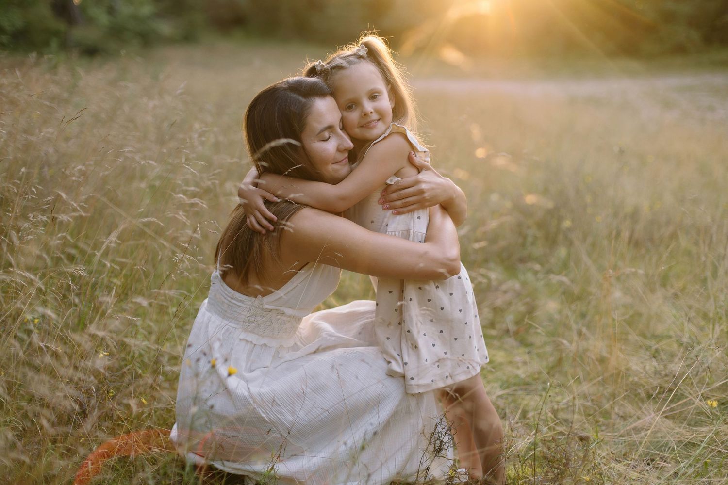 Mother kneeling in golden field hugging young daughter in white dress during sunset.