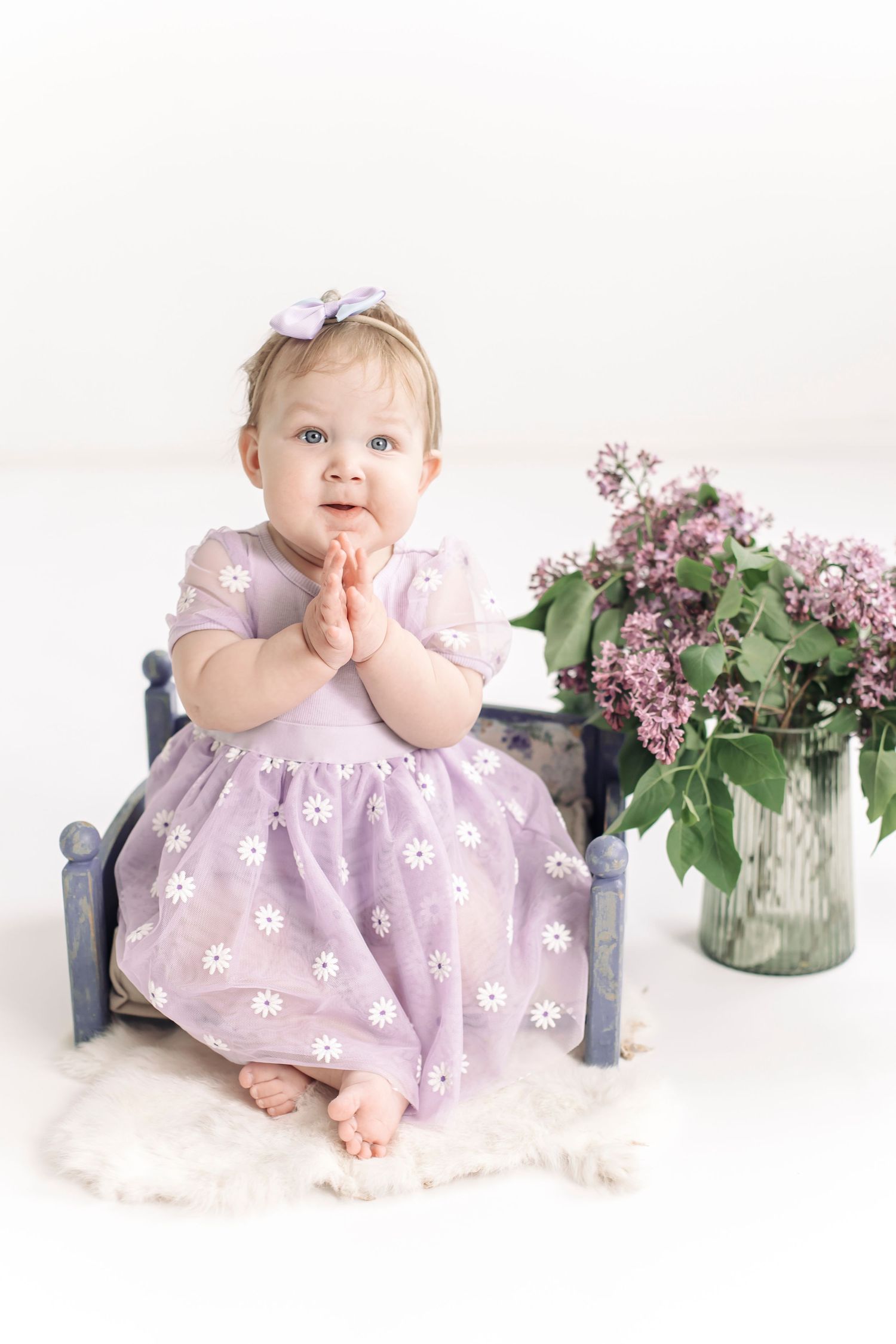 Adorable baby girl in lavender daisy dress clapping hands beside lilac flowers and tiny bed.