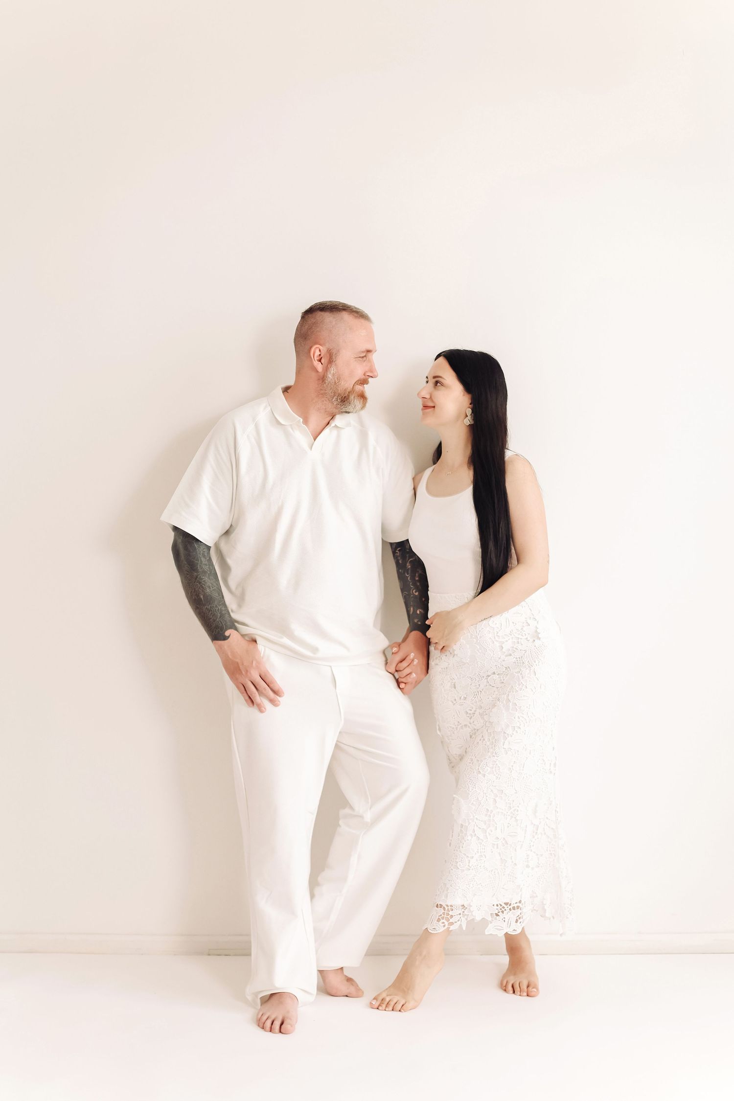 A couple in matching white outfits hold hands and smile at each other against a white background.