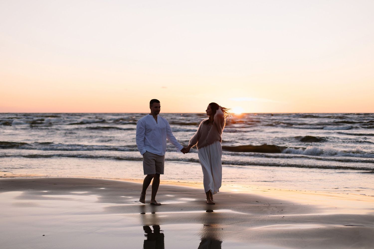 Couple holding hands walking on beach at sunset with ocean waves.