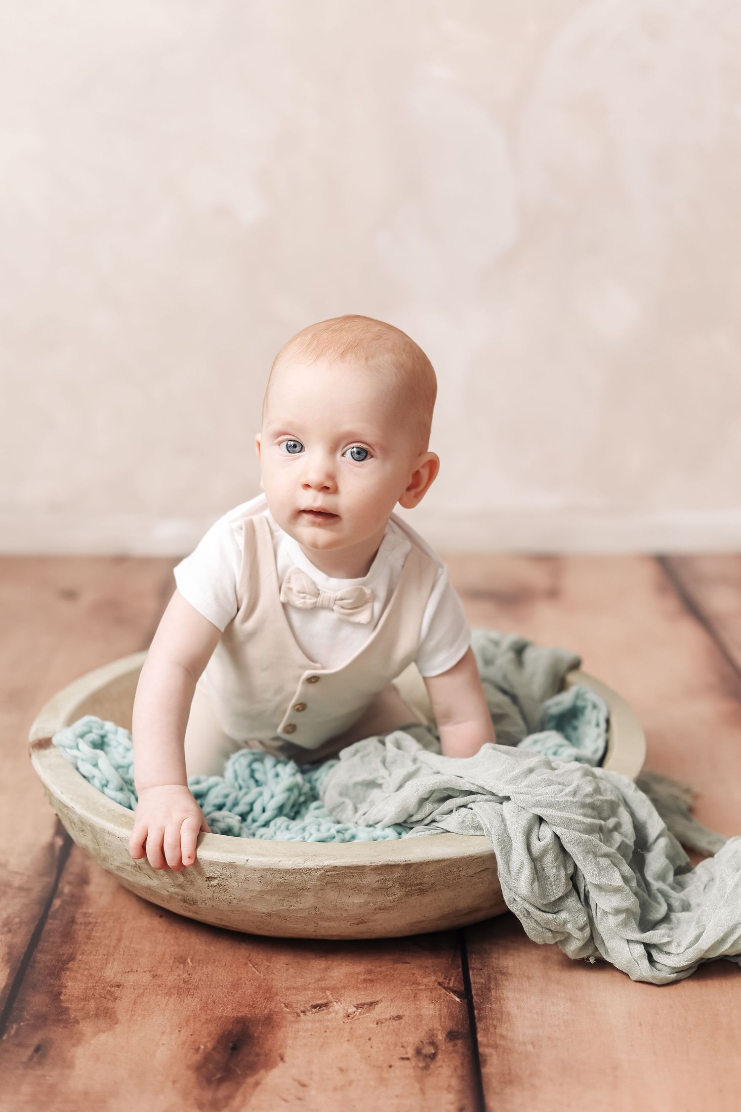 Adorable baby in white bow outfit sitting in wooden bowl with teal blanket on rustic floor.