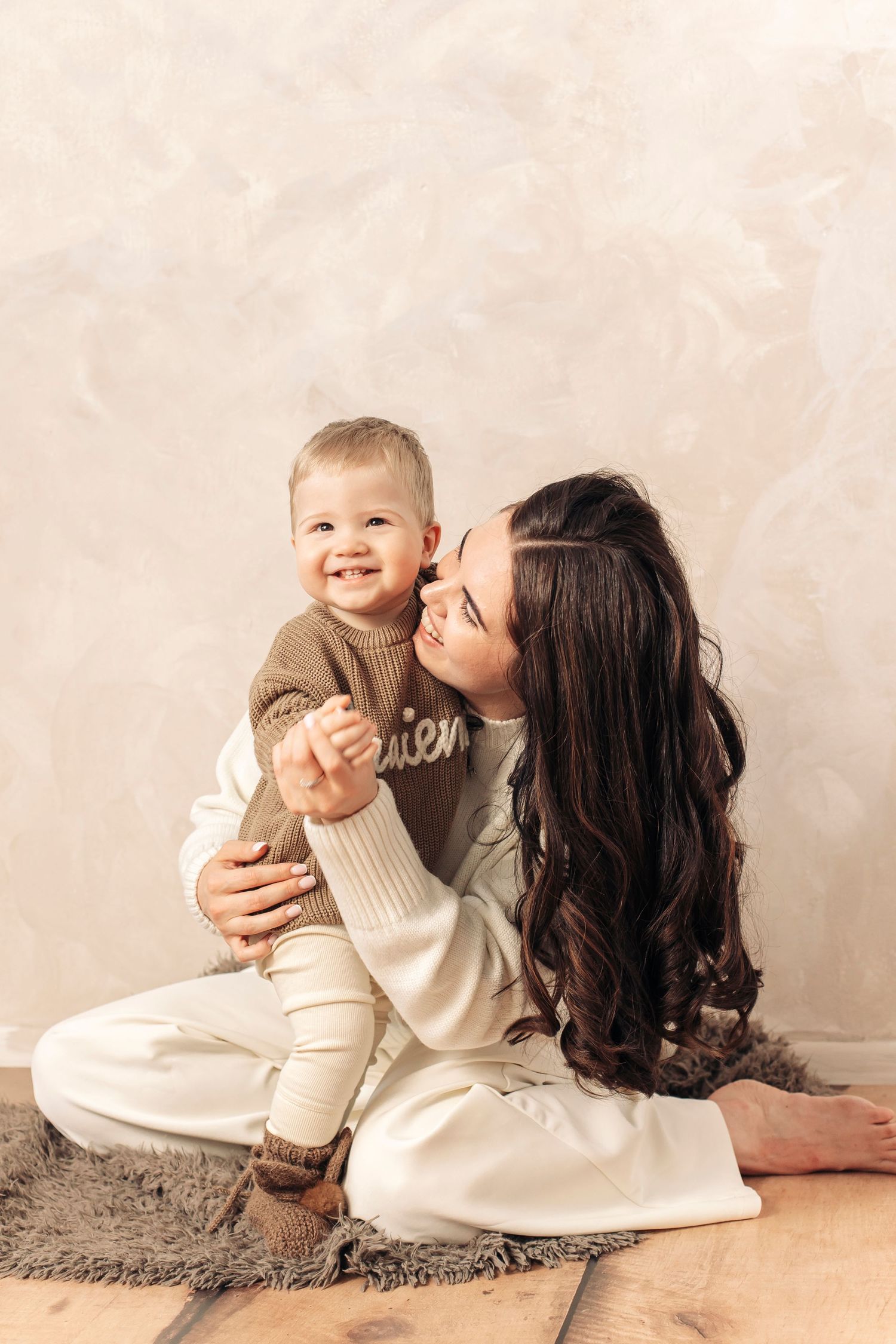 A young mother with long dark hair sits cross-legged on the floor, lovingly holding her smiling baby in a cozy beige sweater.