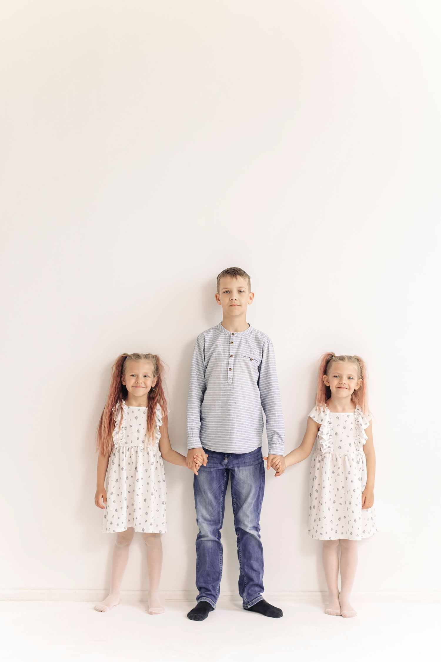 A boy in jeans holds hands with two young girls in matching white floral dresses against a white wall.
