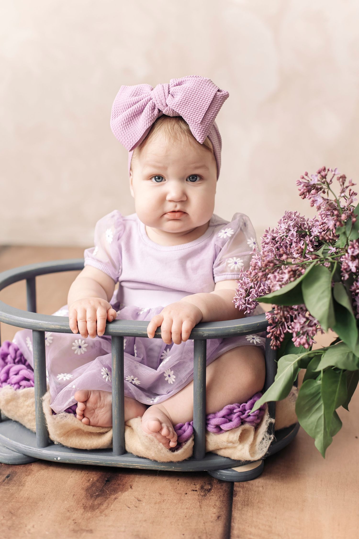 Baby girl in purple floral dress and bow headband sitting in a miniature crib with lilac flowers.