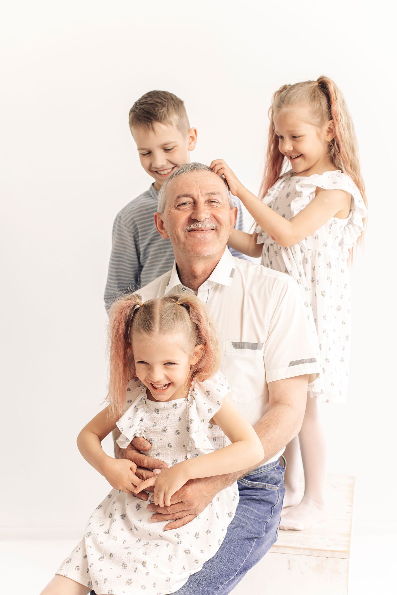 Smiling grandfather with three happy grandchildren playing with his hair in a bright white studio setting.
