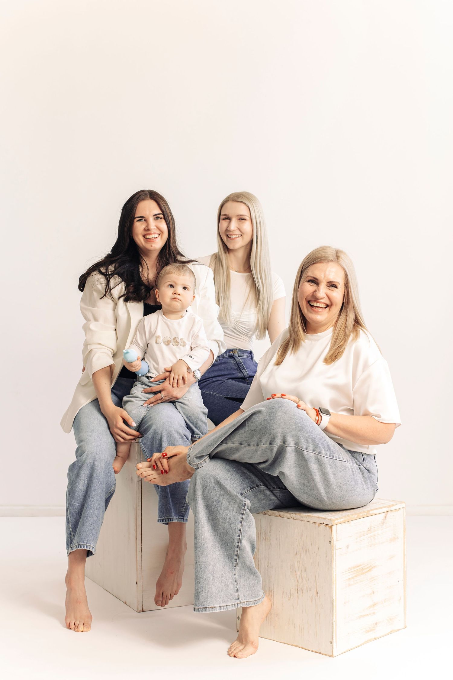 Three generations of women in white tops and jeans pose with a baby on a white wooden box in a bright studio.