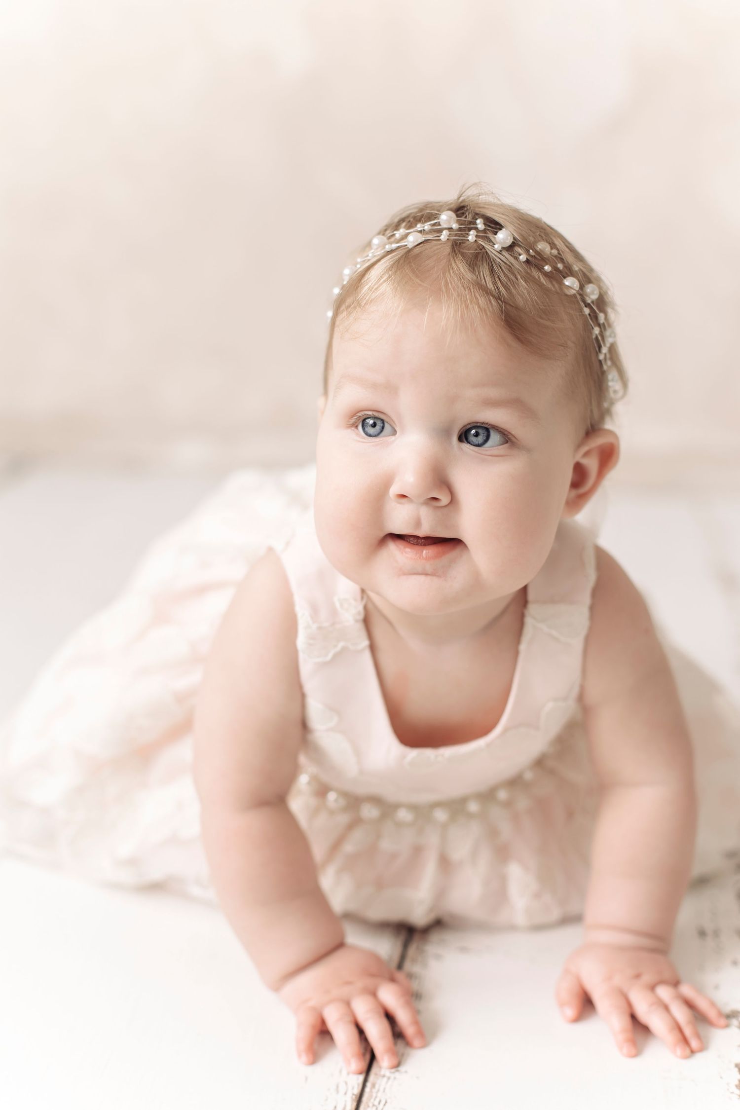 Sweet baby girl in white pearl romper and delicate headband looking up while crawling on floor.
