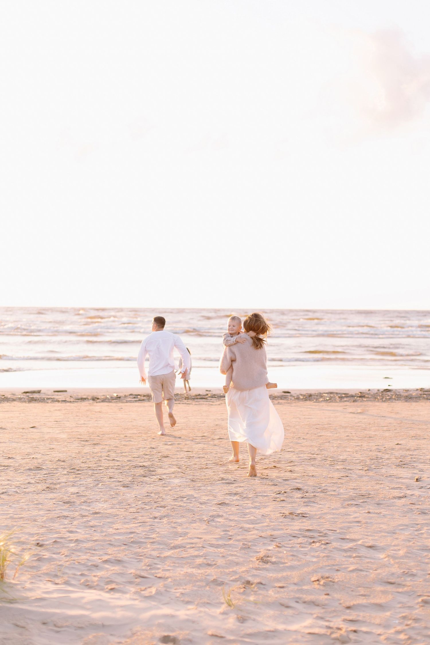 Family running barefoot on sandy beach at sunset, mother carrying baby, father ahead near ocean waves.