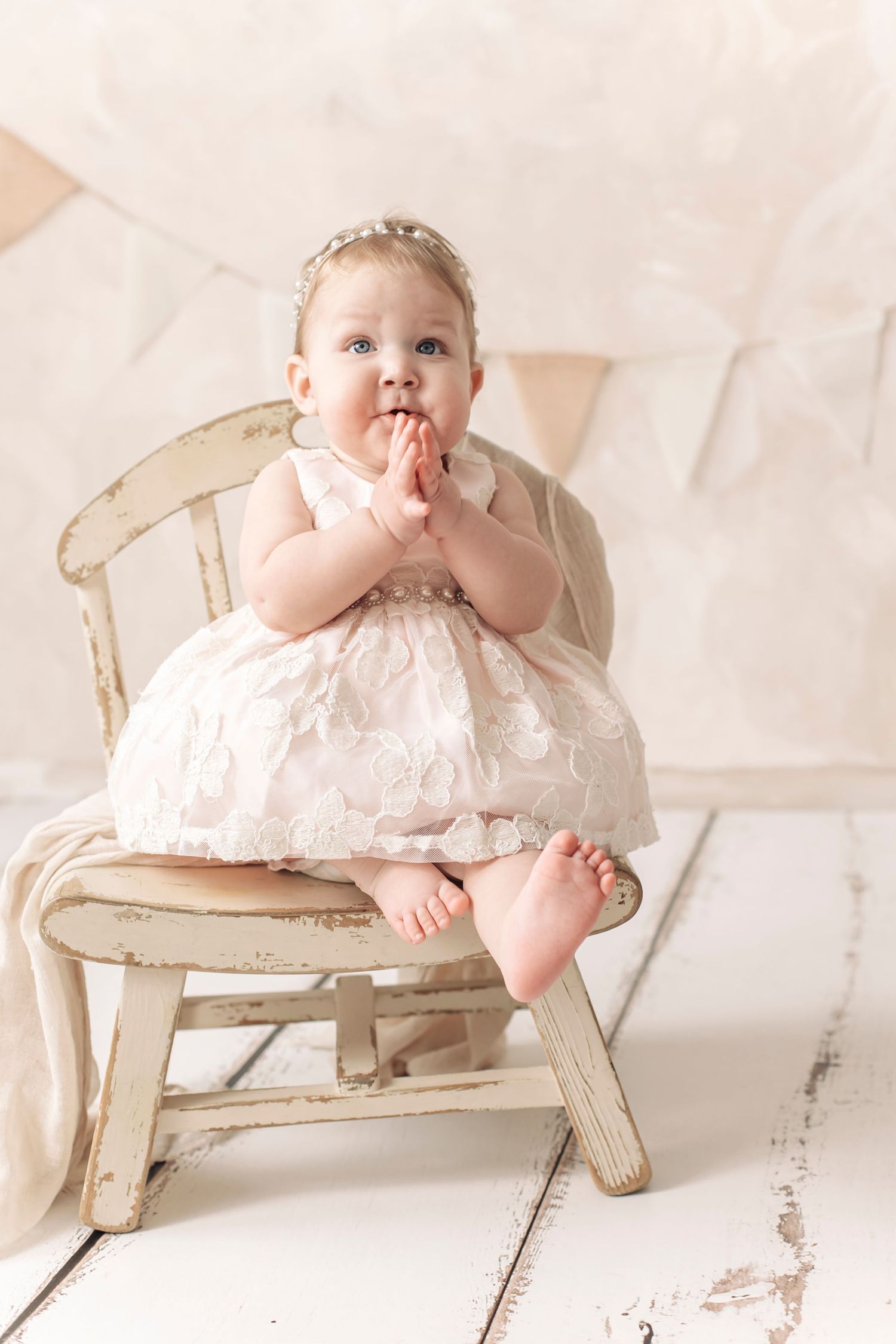 Adorable baby girl in white lace dress sitting on vintage chair with hands together.