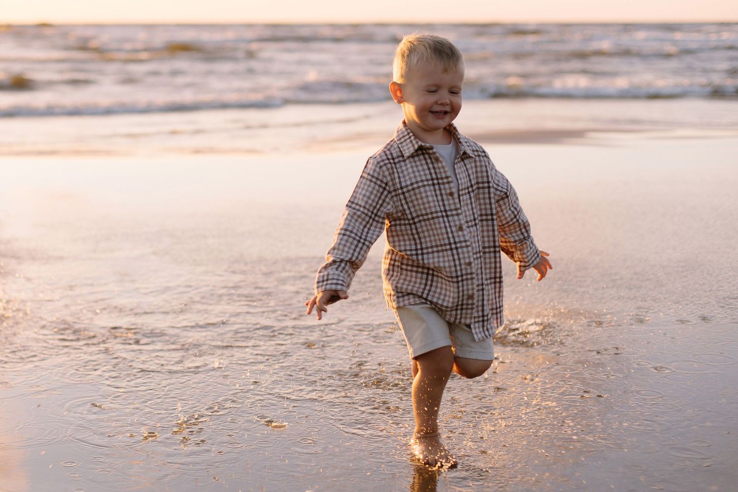 A happy toddler boy runs along a sandy beach at golden sunset, splashing in shallow ocean waves.