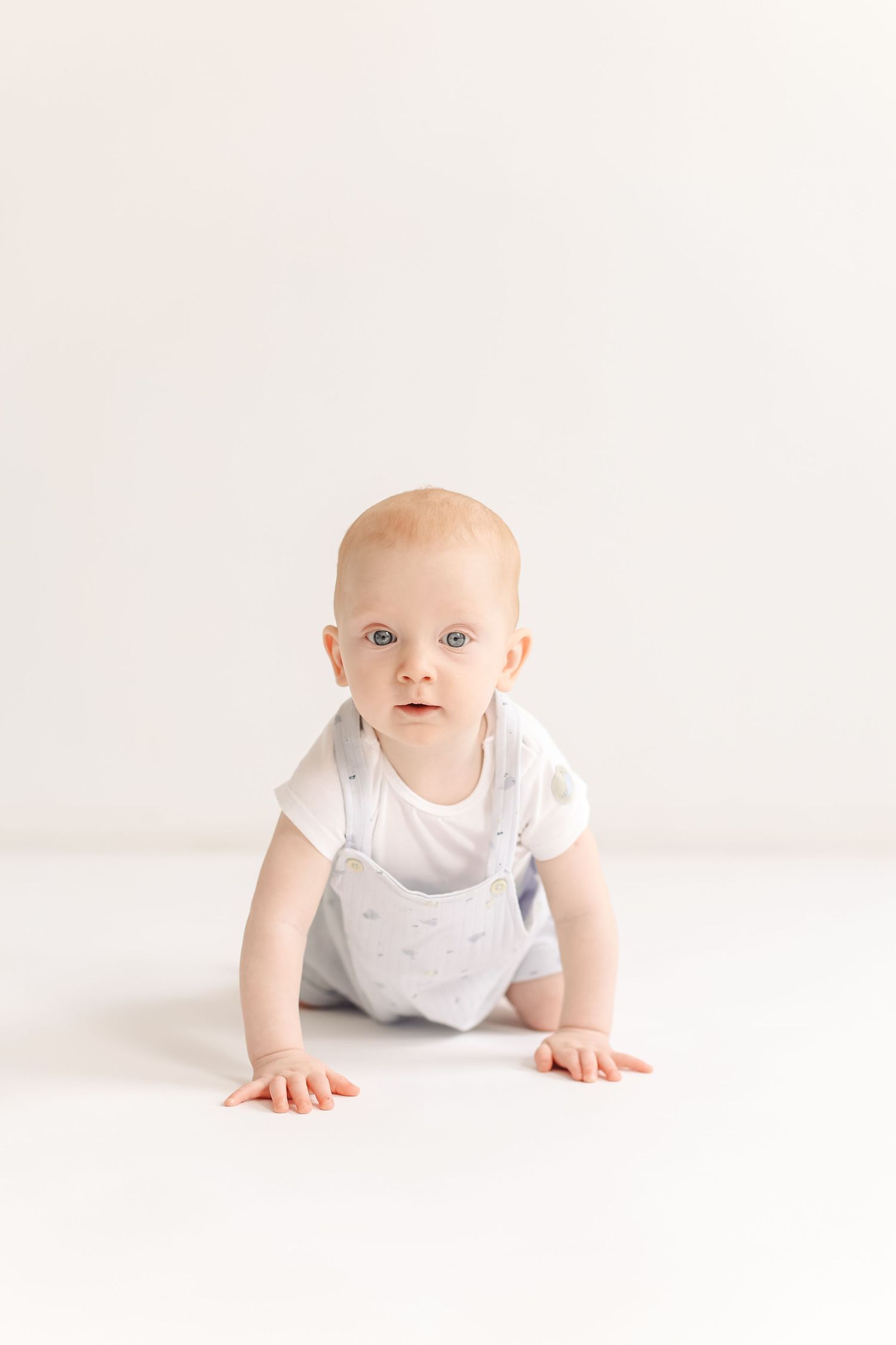 A curious baby in white onesie and gray overalls crawls on a white floor, looking directly at the camera.