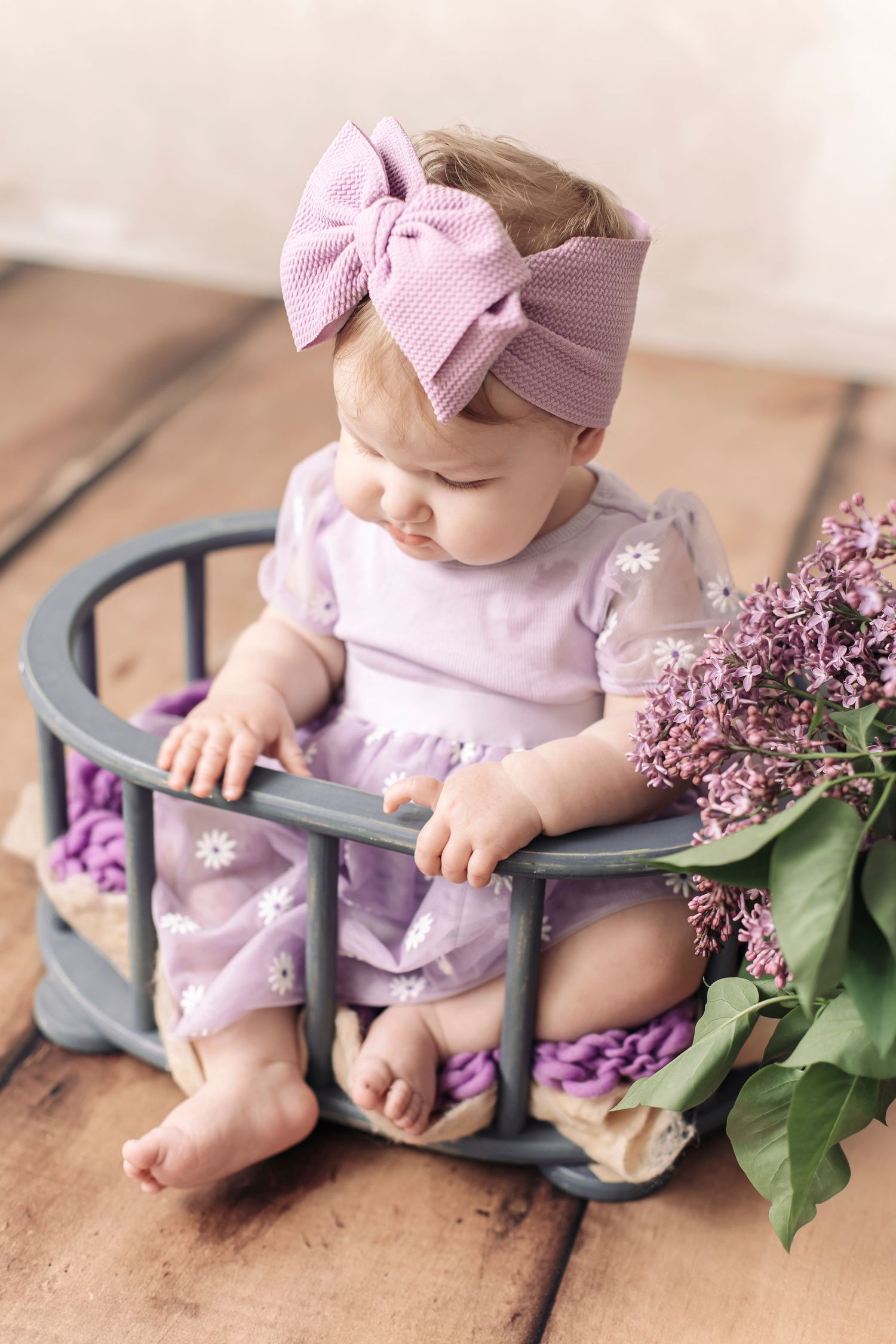 Baby girl in lavender dress and pink bow headband sitting in gray wooden crate with lilac flowers.