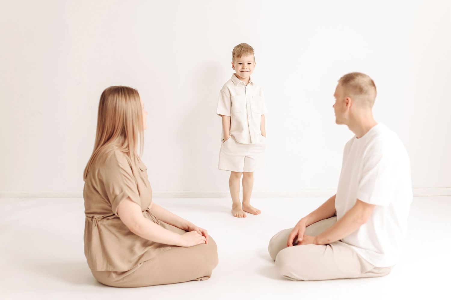Family of three in minimalist white room, parents sitting cross-legged on floor while young boy stands between them.