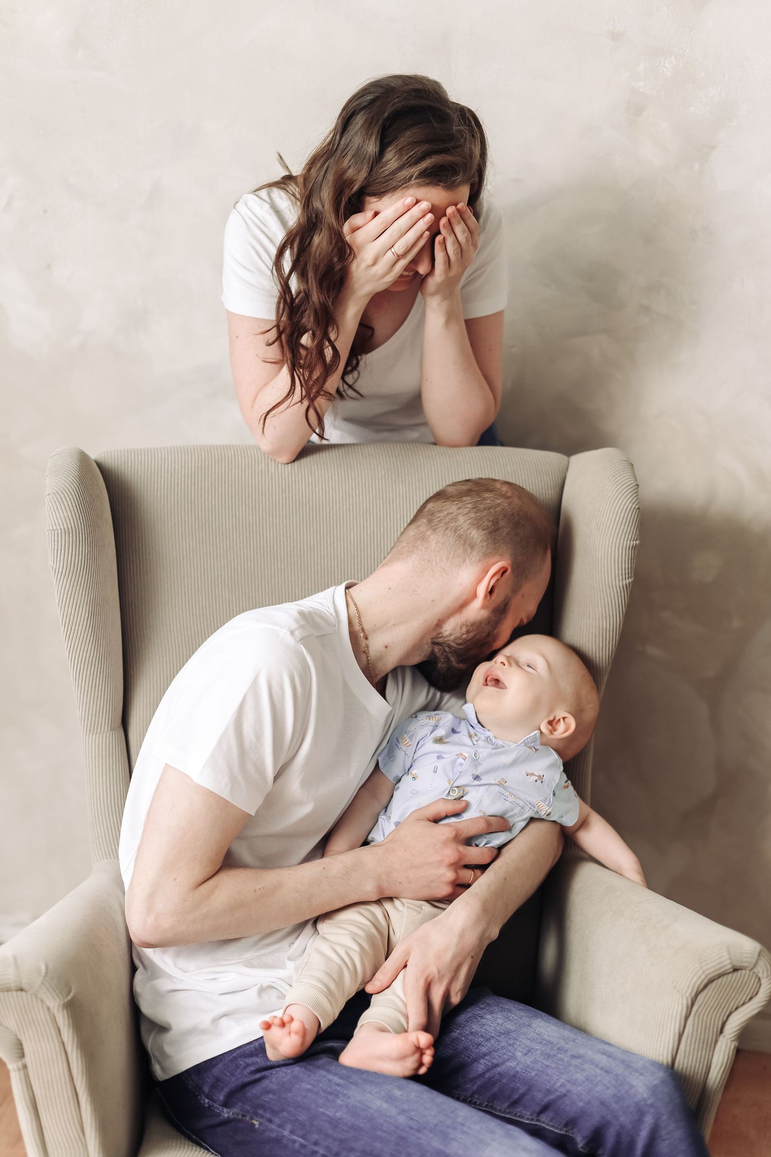 Father holding crying baby in chair while exhausted mother covers her face, depicting postpartum stress.