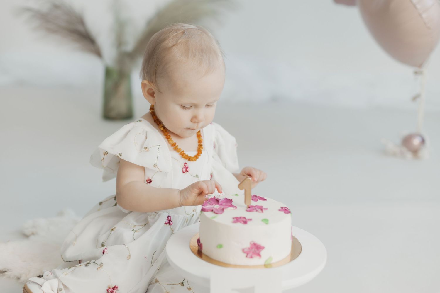 Baby girl in white dress touches her first birthday cake decorated with pink stars.Cake smash fotosesija Rīgā studijā, mazulis ar kūku.