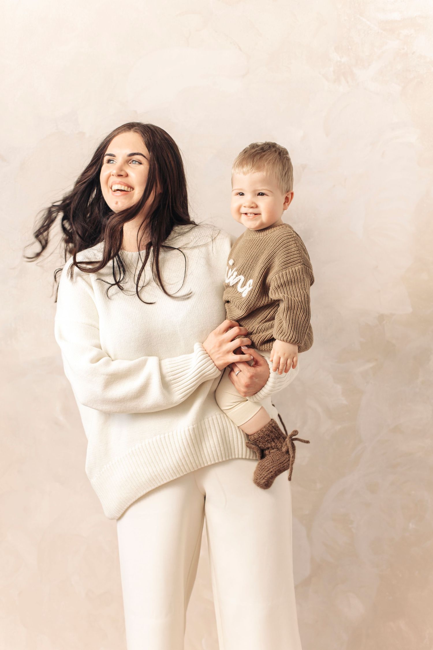 A smiling young mother in white outfit holds her happy toddler in a brown knit sweater against a beige wall.