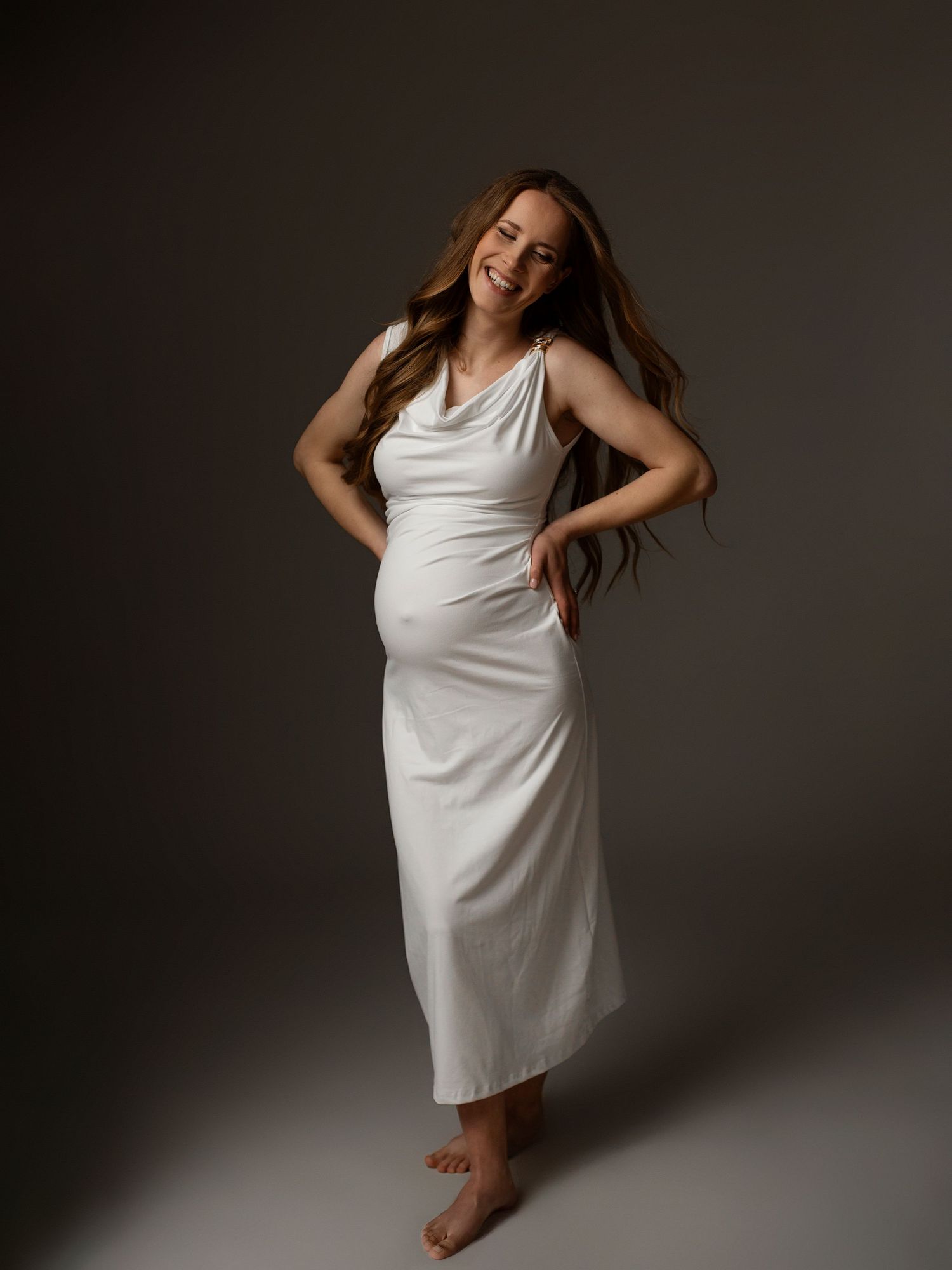 Smiling pregnant woman in white maxi dress posing barefoot against gray studio background.Gaidību fotosesija Rīgā- grūtnieču fotosesija Latv