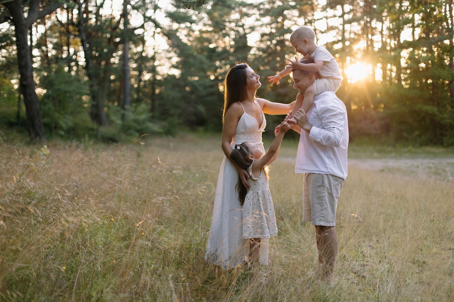 Family enjoying golden sunset in a meadow with pine trees, parents playing with two young children outdoors.