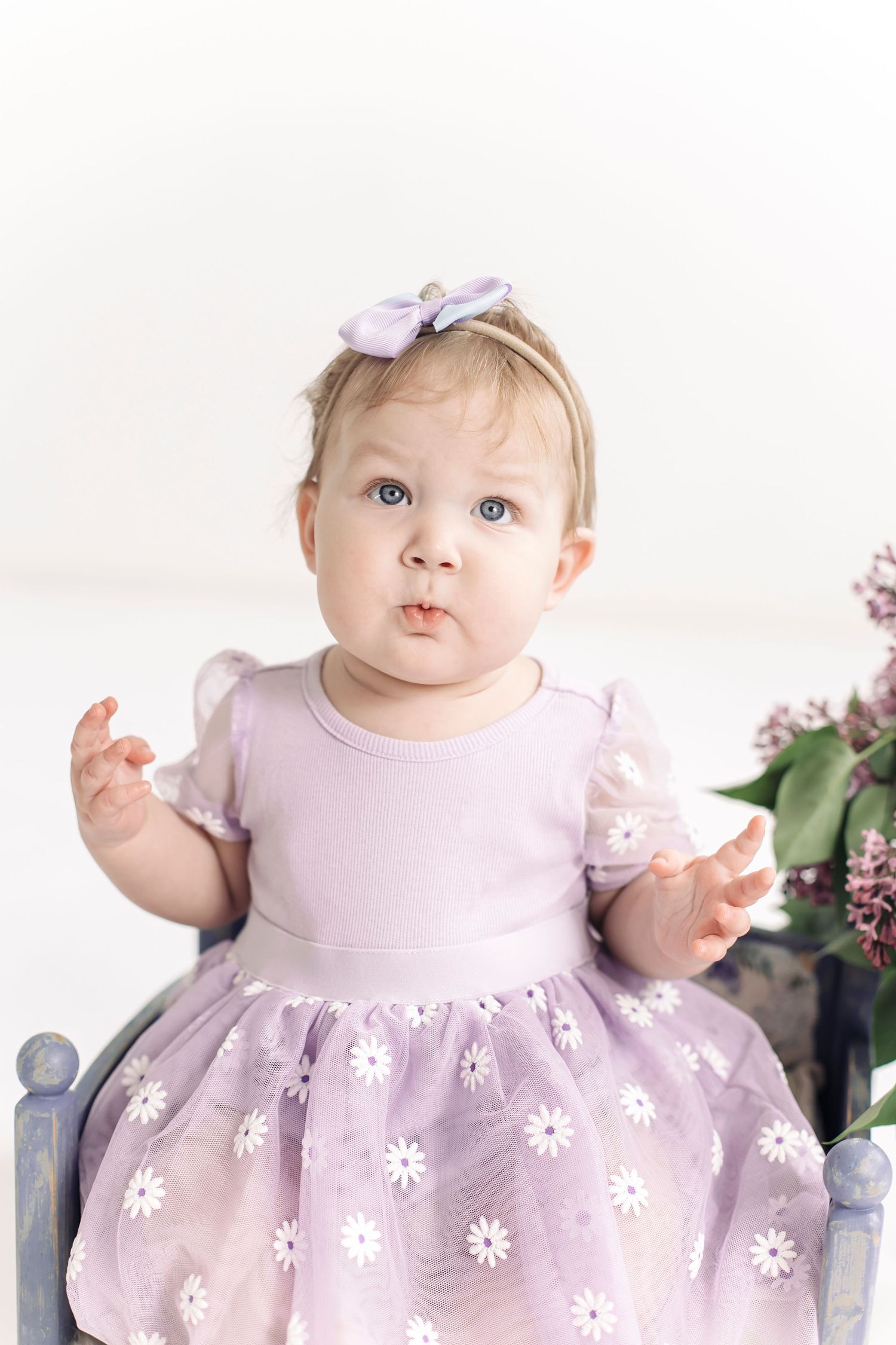 Adorable baby girl in purple floral dress with bow headband sits gracefully, blue eyes wide with curiosity.