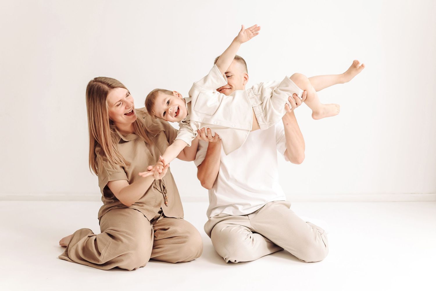 Happy family sitting on white floor, parents playfully lifting laughing baby in beige casual outfits.