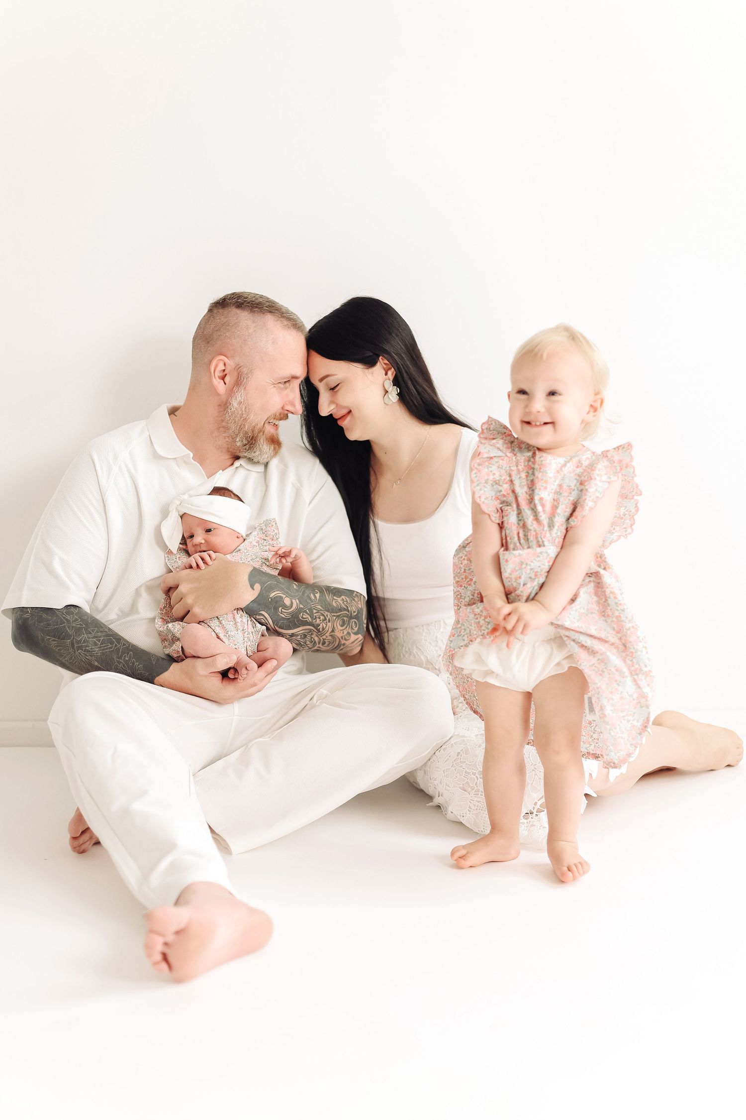 A tattooed father and dark-haired mother sit on white floor holding newborn baby while toddler daughter in floral dress smiles beside them.