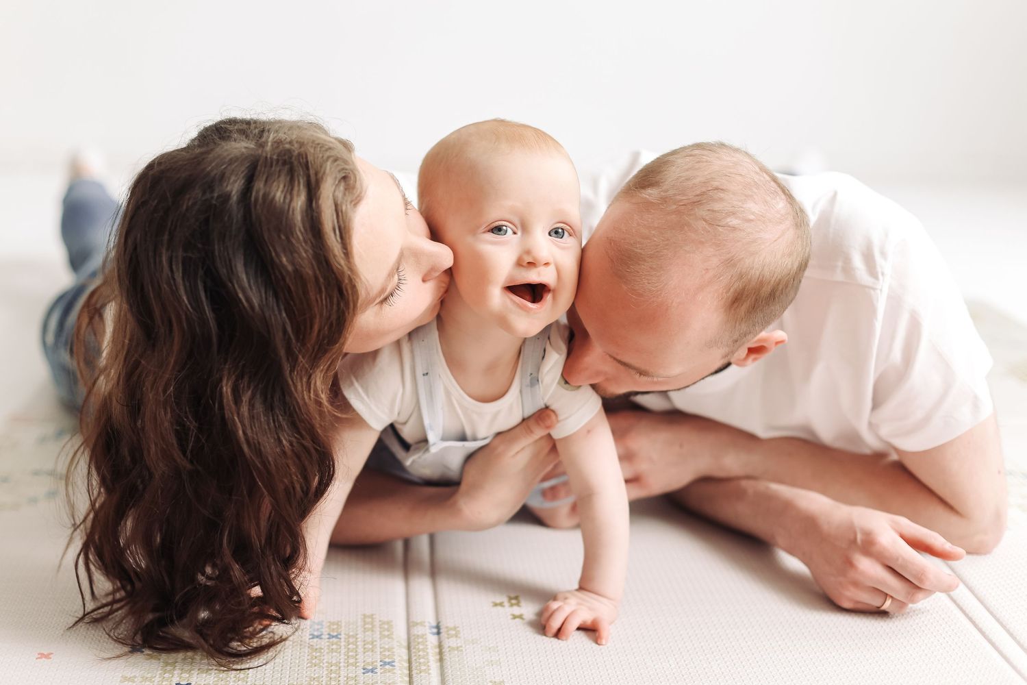 Happy baby laughing as loving parents kiss cheeks during family photoshoot on white floor.