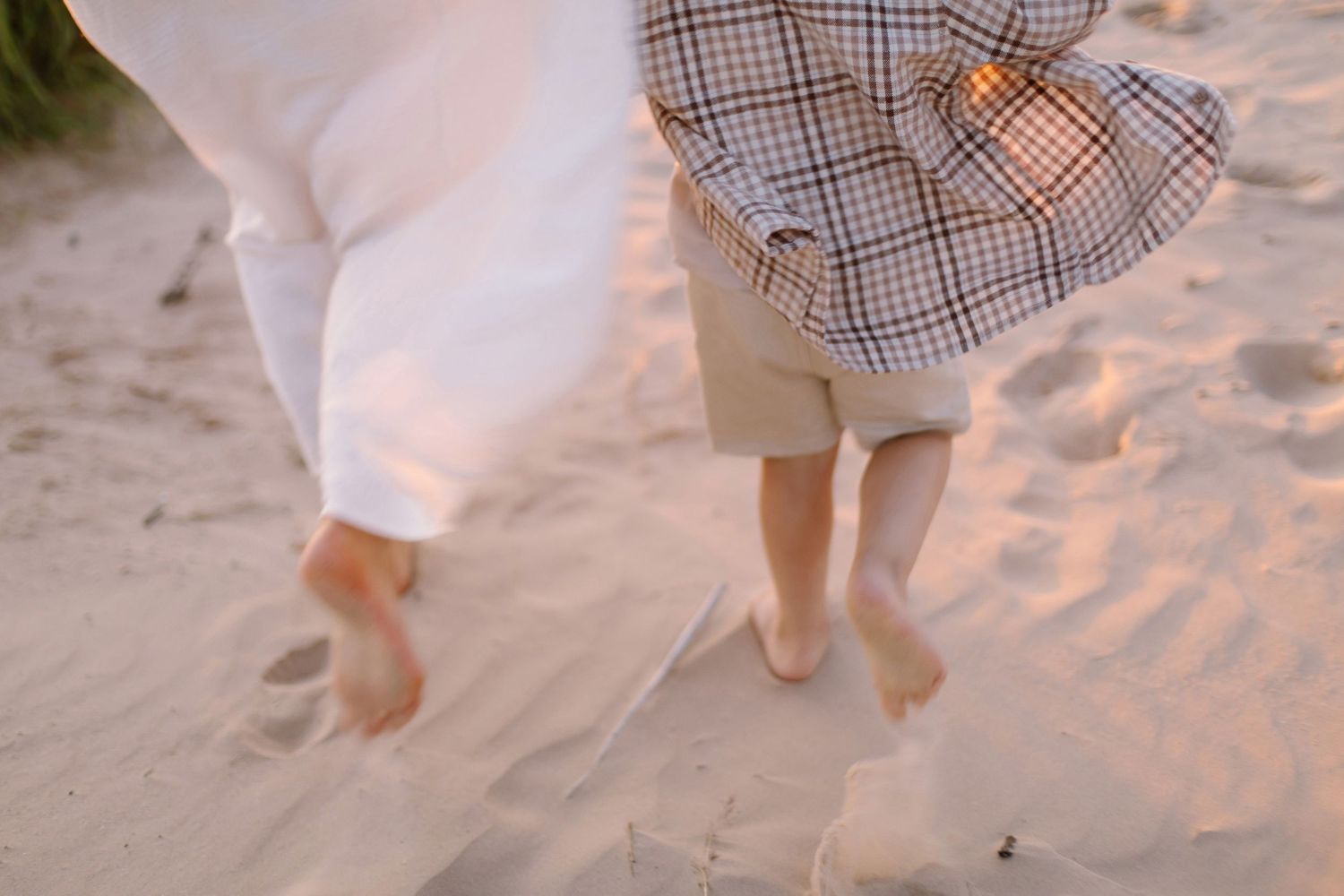 Two barefoot children walking on sandy beach, one in white dress and other in plaid shirt and tan shorts.